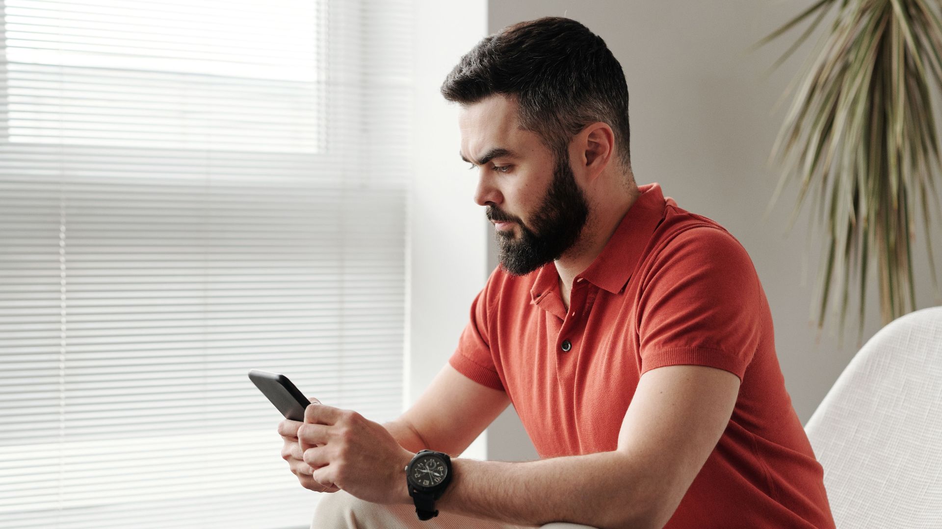 Bearded man in orange shirt text messaging on smartphone while seated indoors.