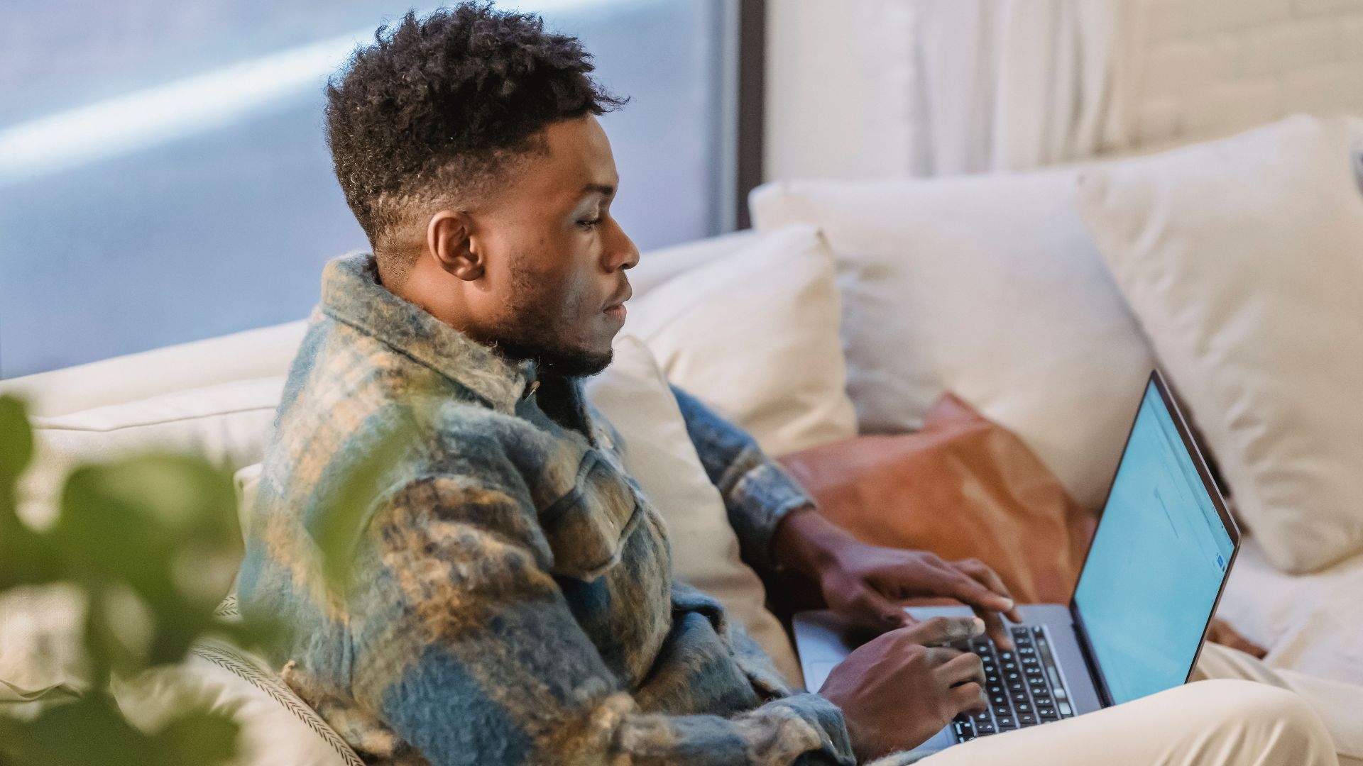 Side view of concentrated African American male sitting on soft couch near window and browsing internet on laptop and looking at screen
