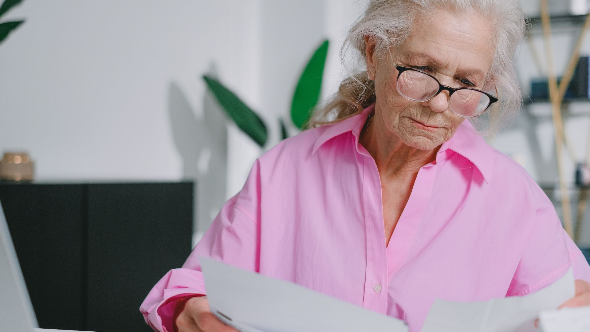 Elderly woman with eyeglasses reading papers at a desk, focusing indoors.