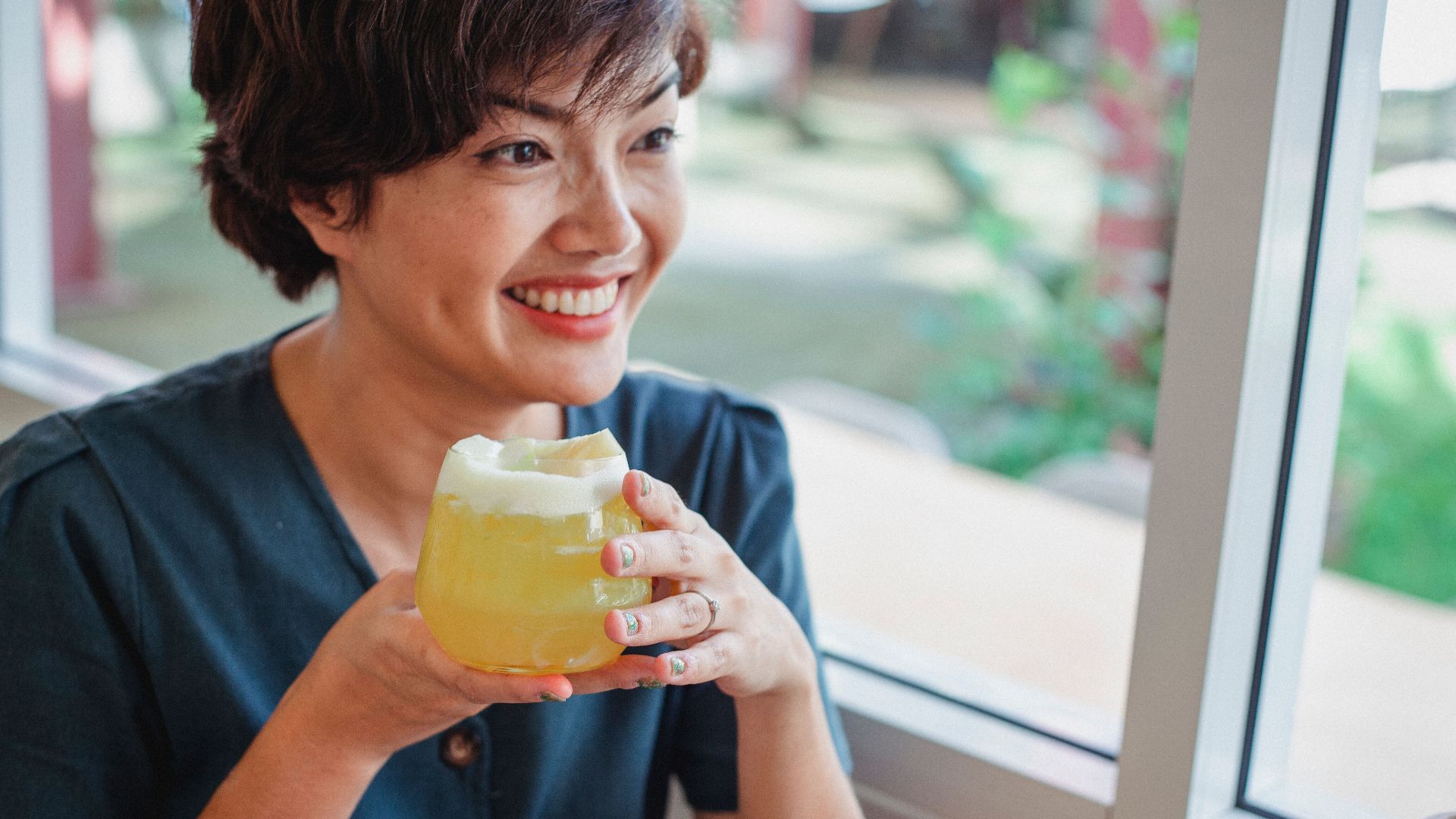 Positive Asian female smiling while sitting in restaurant near window with blurred street with glass of yellow aperitif with foam