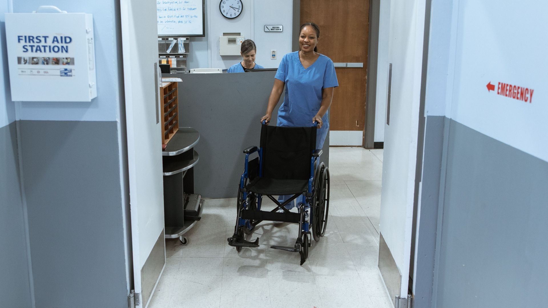 A nurse in scrubs pushes an empty wheelchair through a hospital hallway.