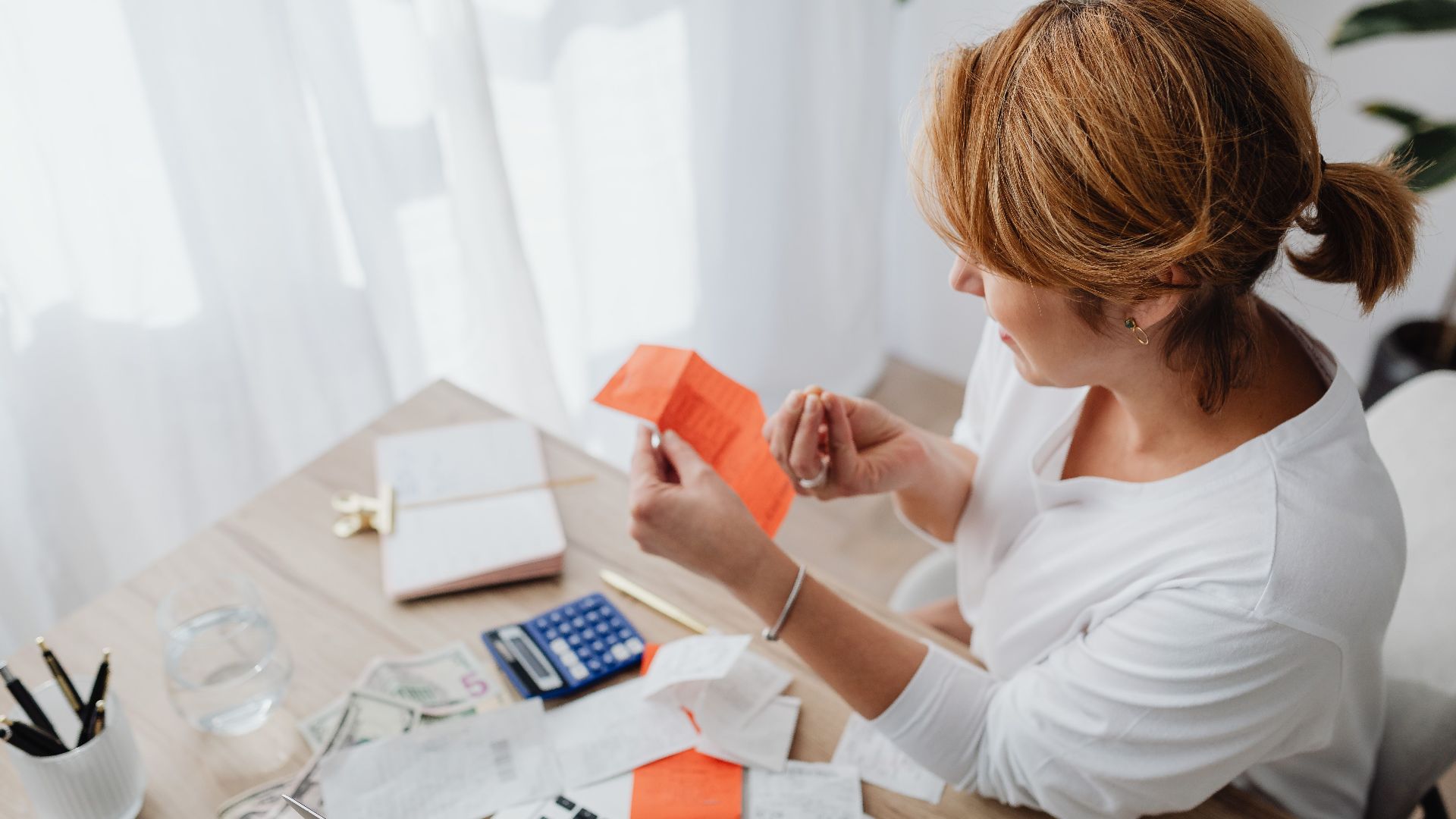 A woman organizing her finances with bills and receipts at a home office desk using a calculator and laptop.