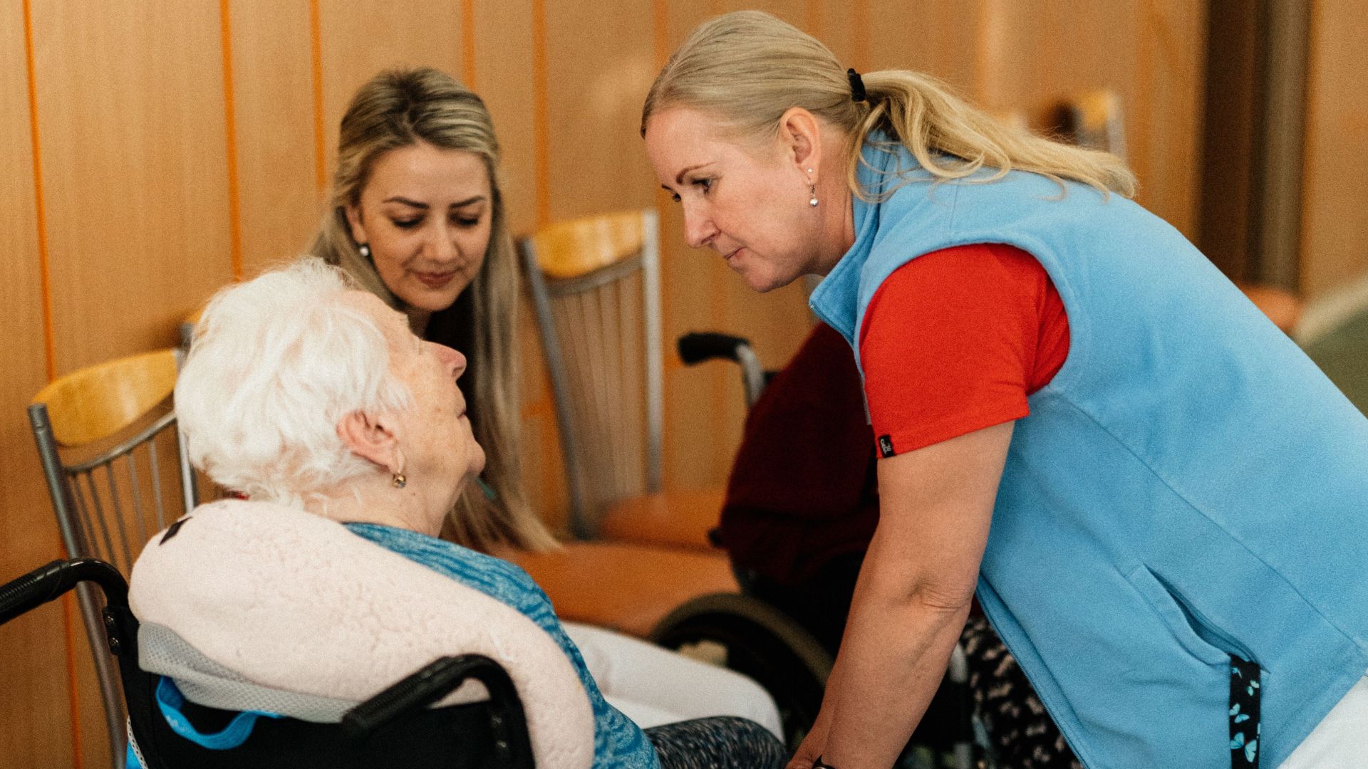 A caregiver talks with elderly women in a wheelchair at a retirement home.