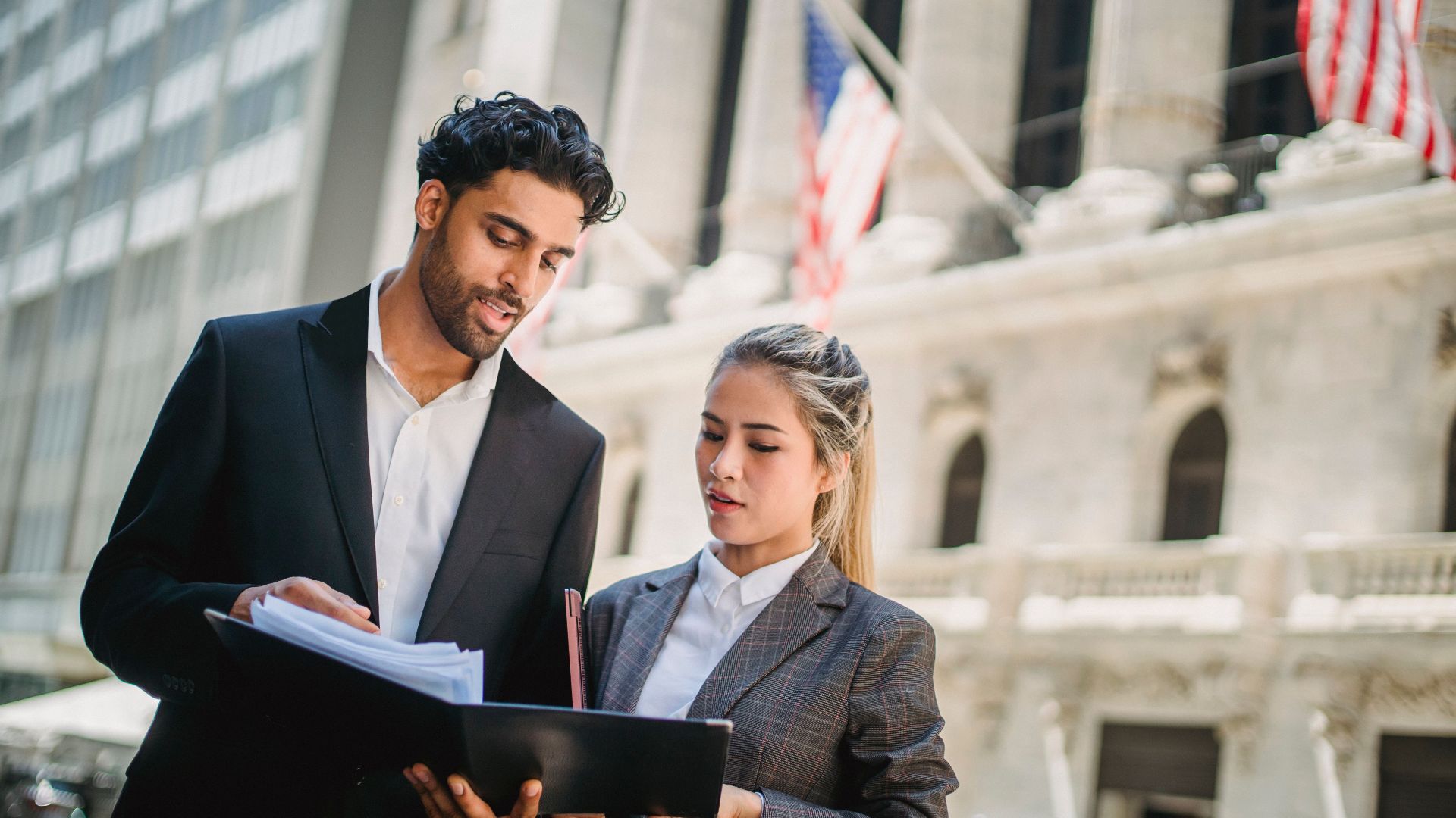 Two business professionals review documents outside a financial institution, indicating teamwork and collaboration.