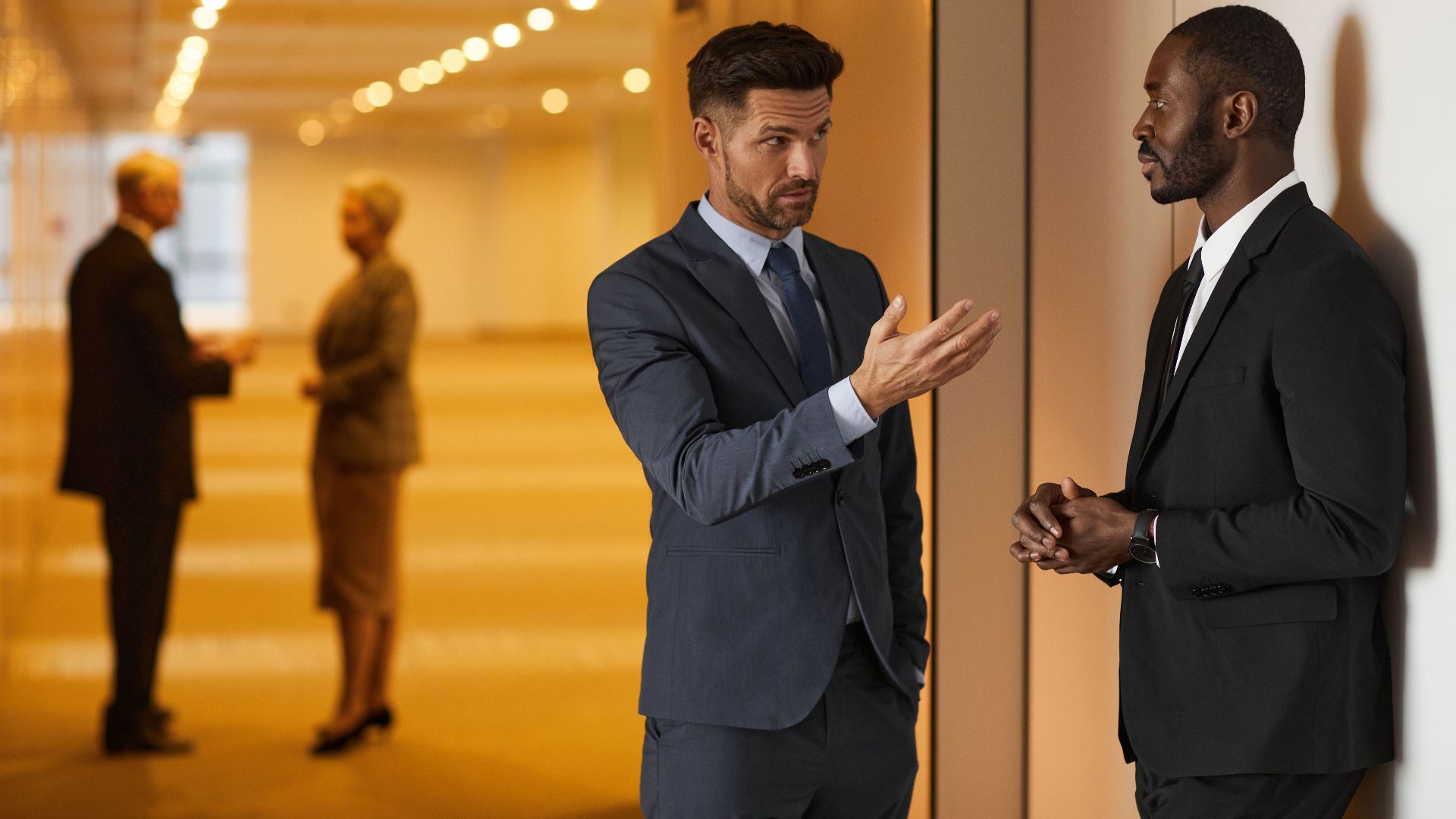 Two businessmen in formal attire engaged in a serious discussion in a modern office hallway.