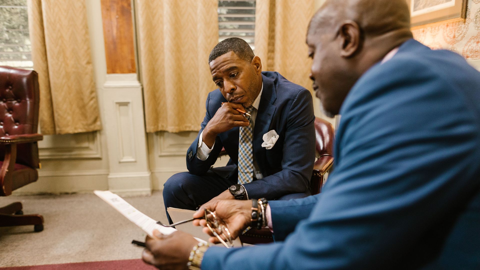 Two executives in business attire discuss documents in an office setting.