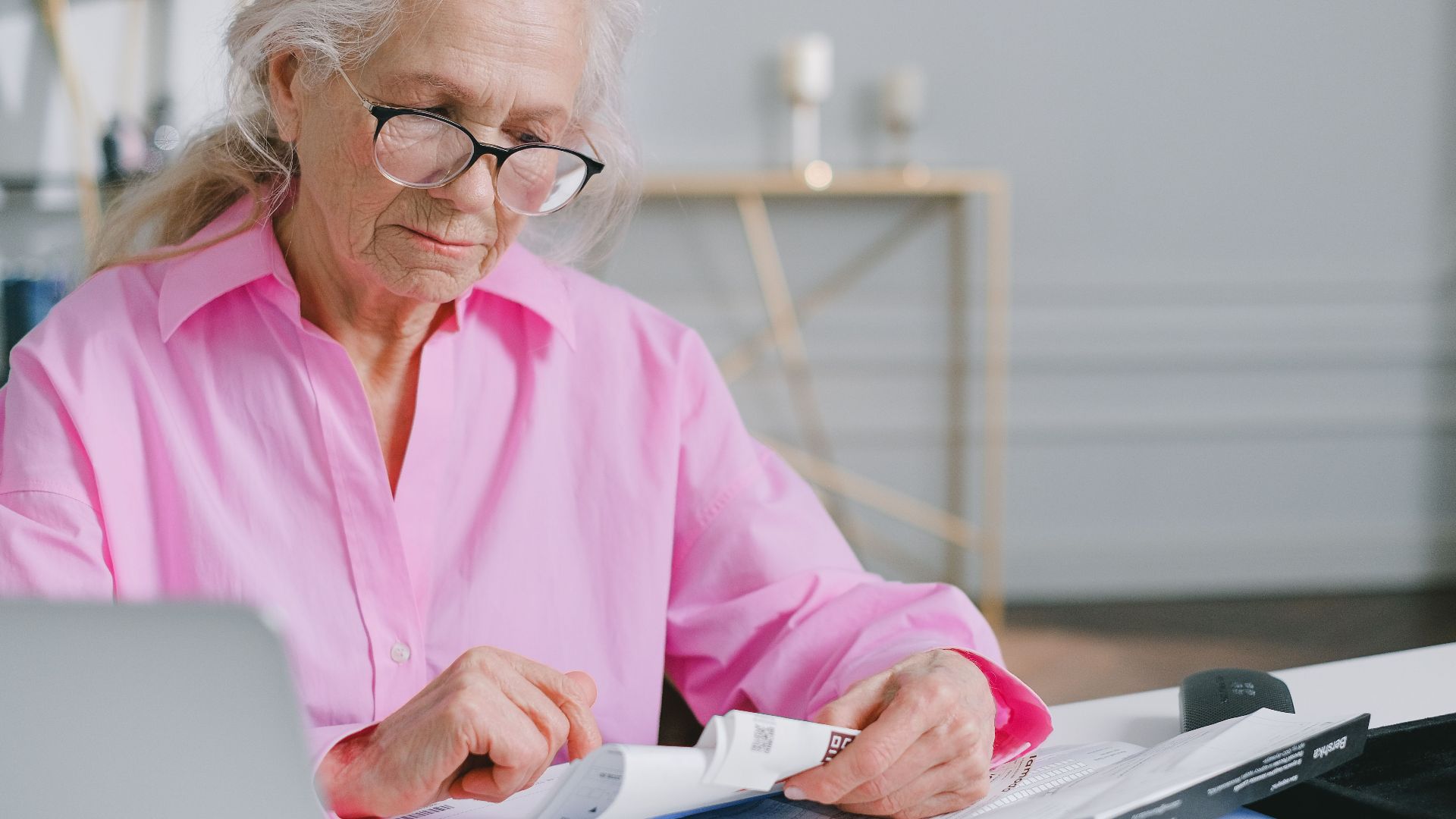 Elderly woman in pink shirt reading documents at a desk indoors.