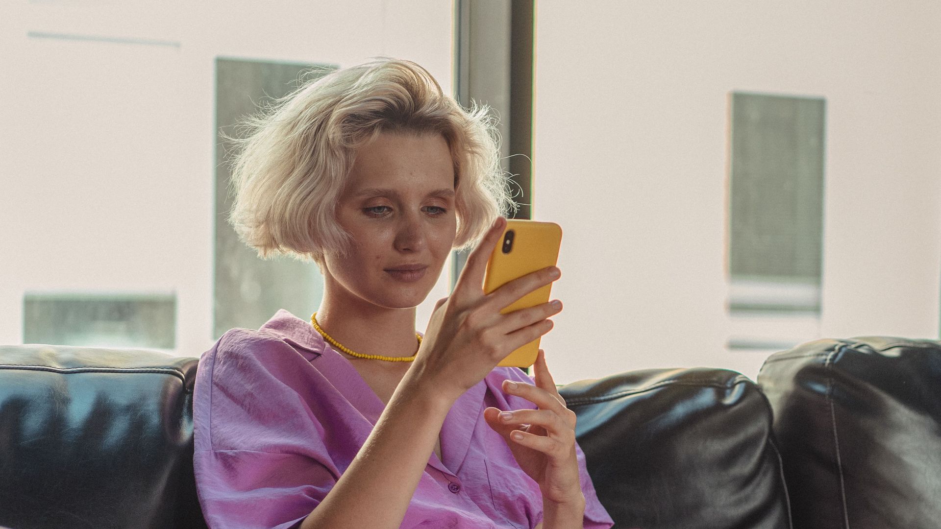 Woman in pink shirt using smartphone on a leather couch in a bright room.