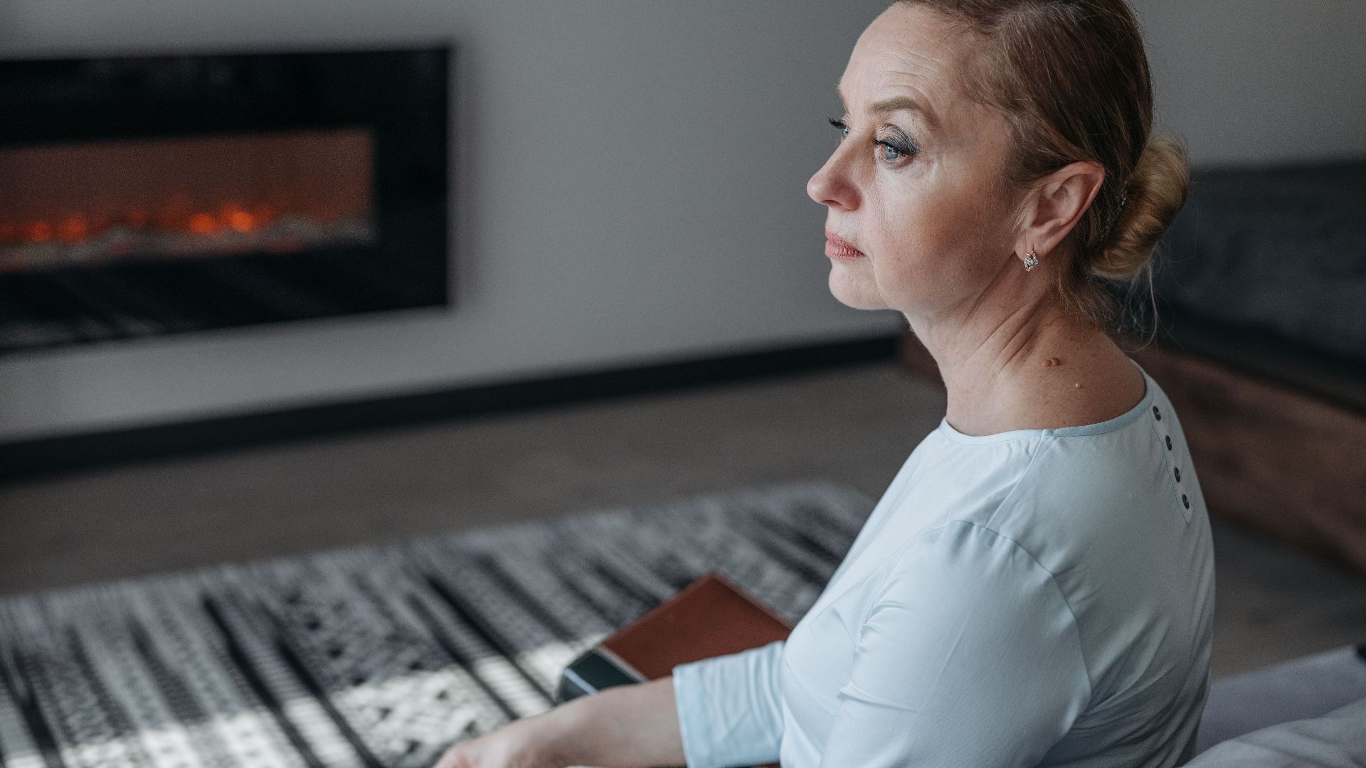 Senior woman sitting indoors, reflecting near a warm fireplace, calm atmosphere.