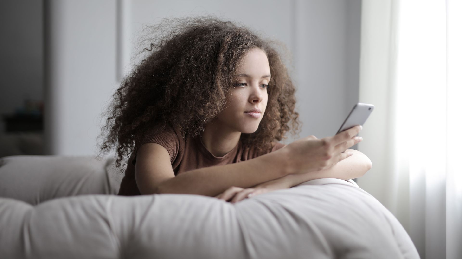 A young woman with curly hair comfortably using her smartphone while sitting indoors.