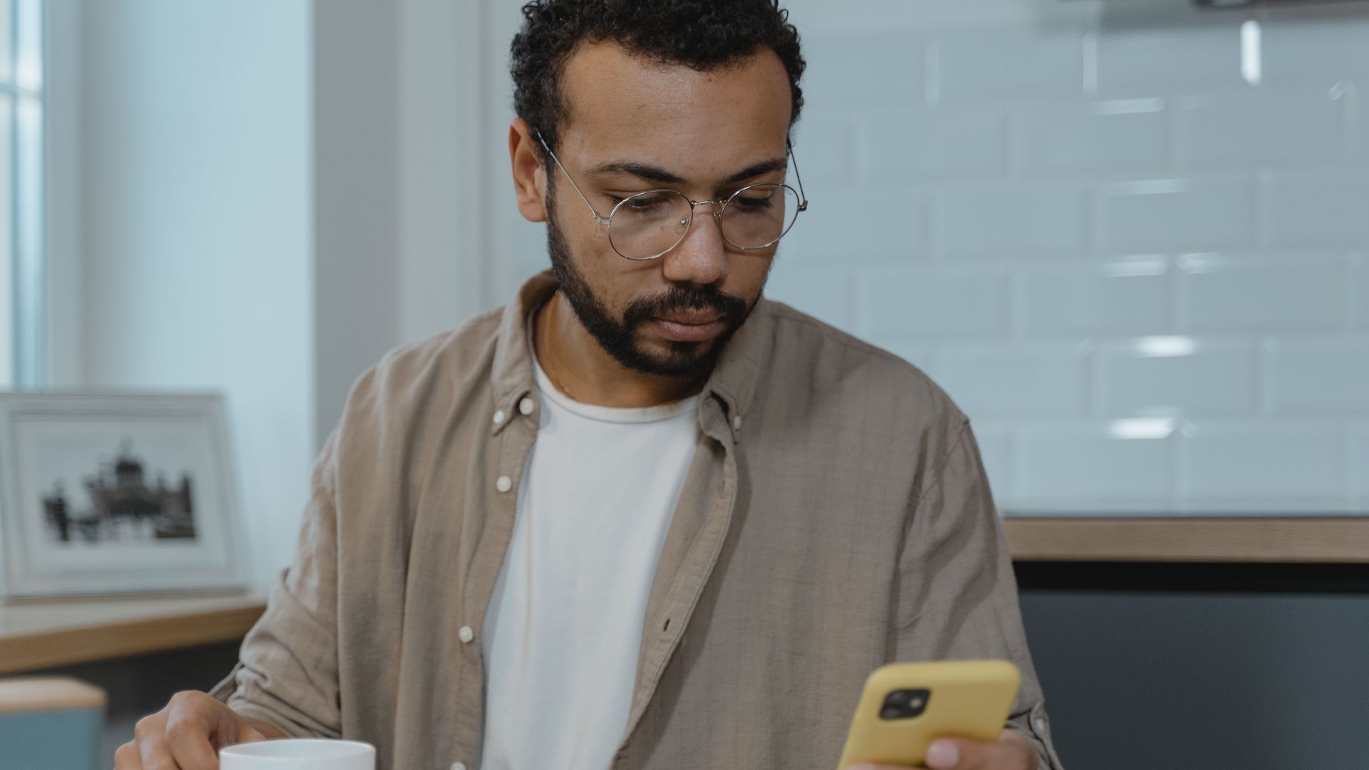 A man with eyeglasses using a smartphone while enjoying breakfast indoors. Perfect for lifestyle and technology themes.