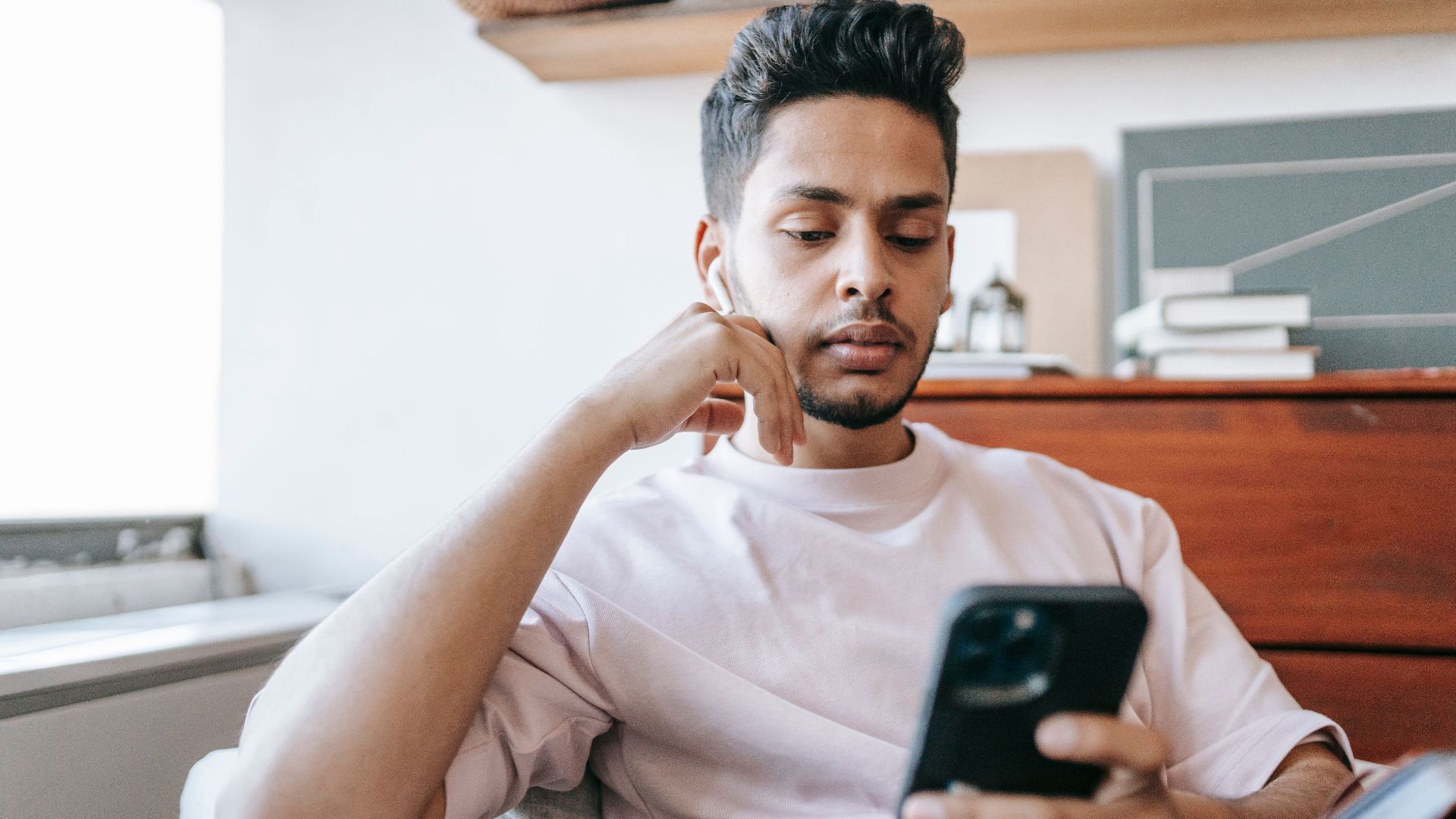 Serious Indian male listening to music in wireless earphones while browsing modern cellphone in armchair in light room at home