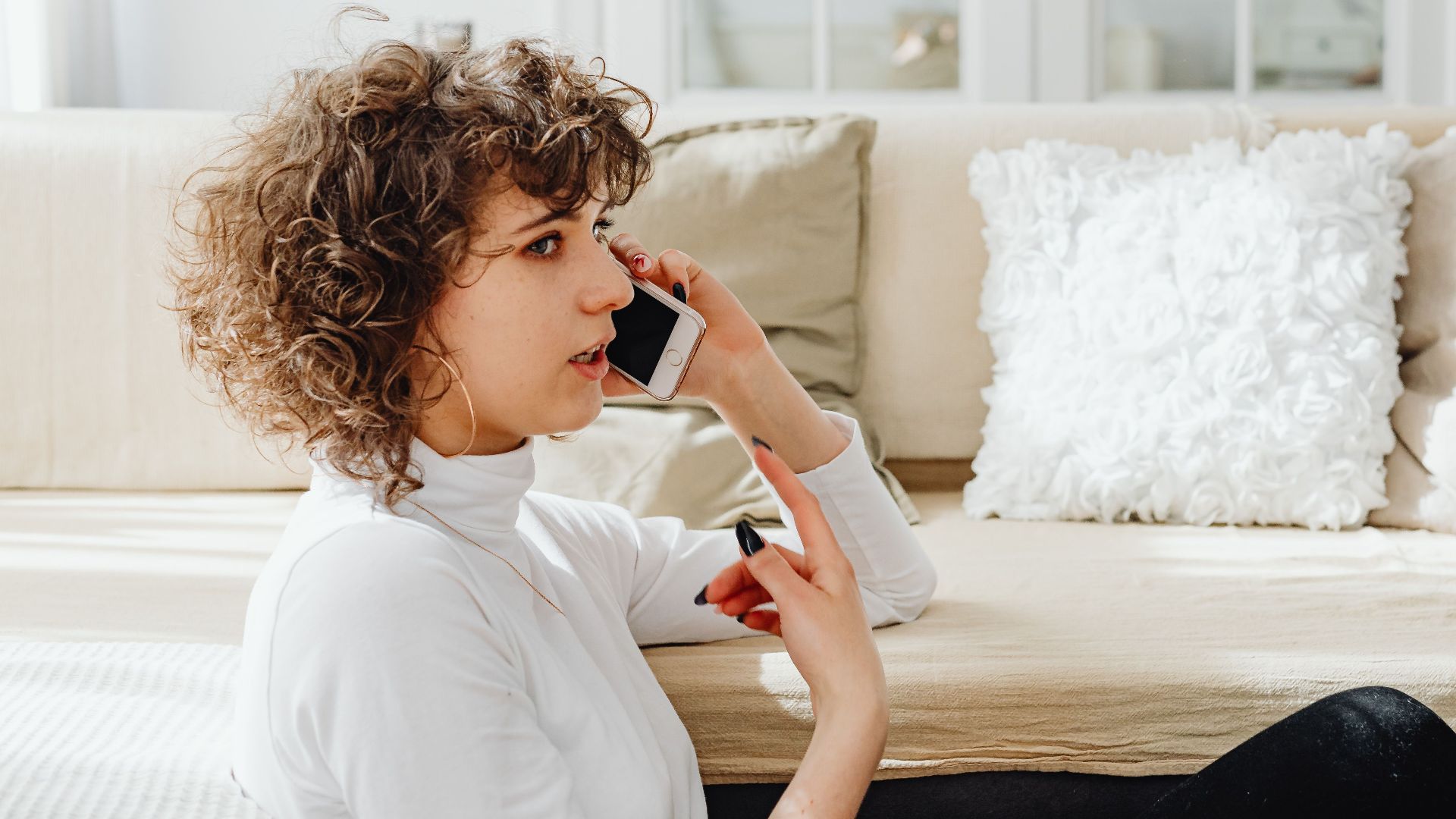 A woman is seated, talking on her phone in a bright, cozy living room.