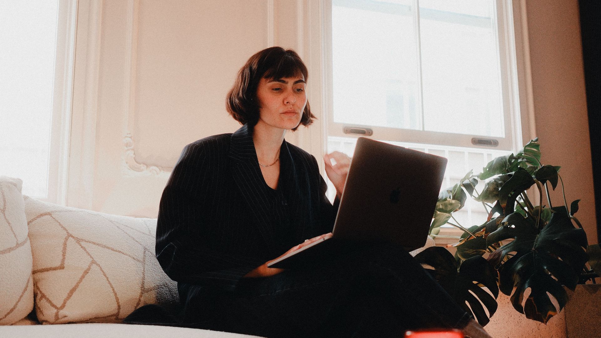 Woman in suit using laptop on sofa in bright room, modern lifestyle.