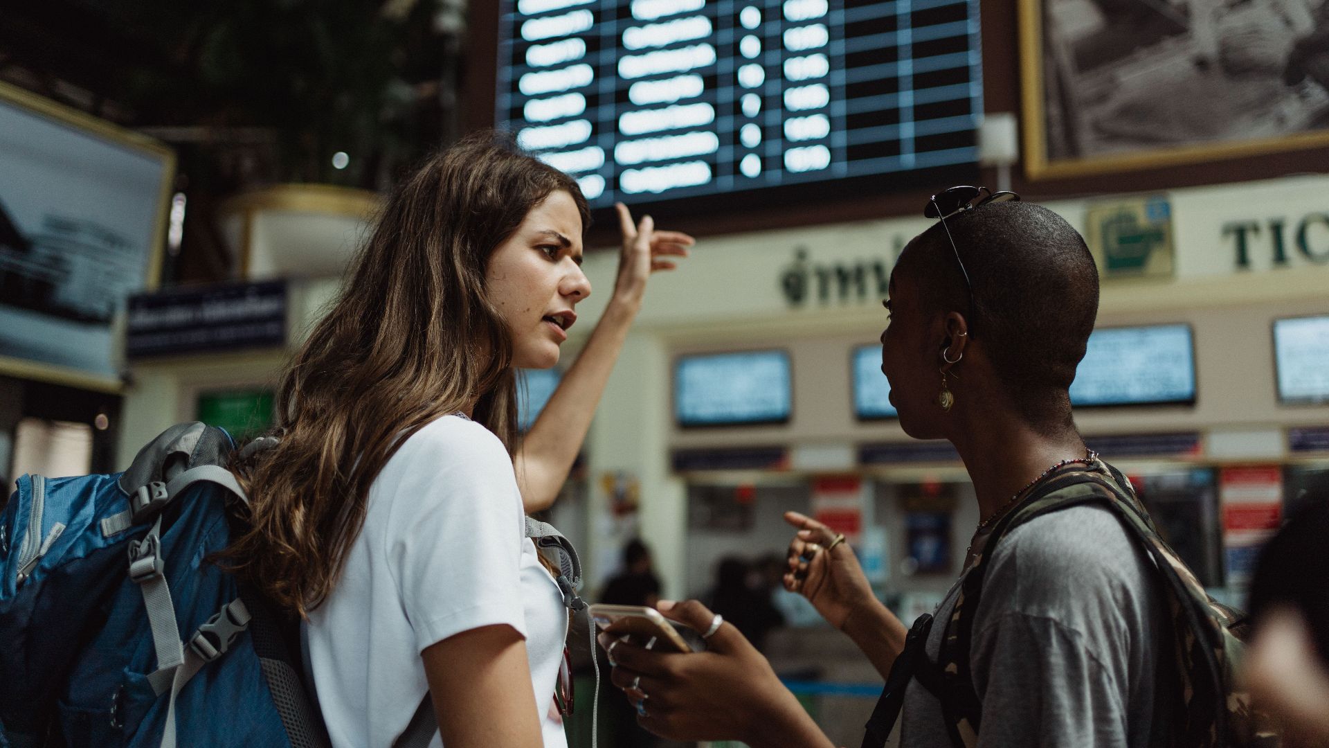 Two travelers with backpacks discuss in front of an airport timetable. Adventure awaits.