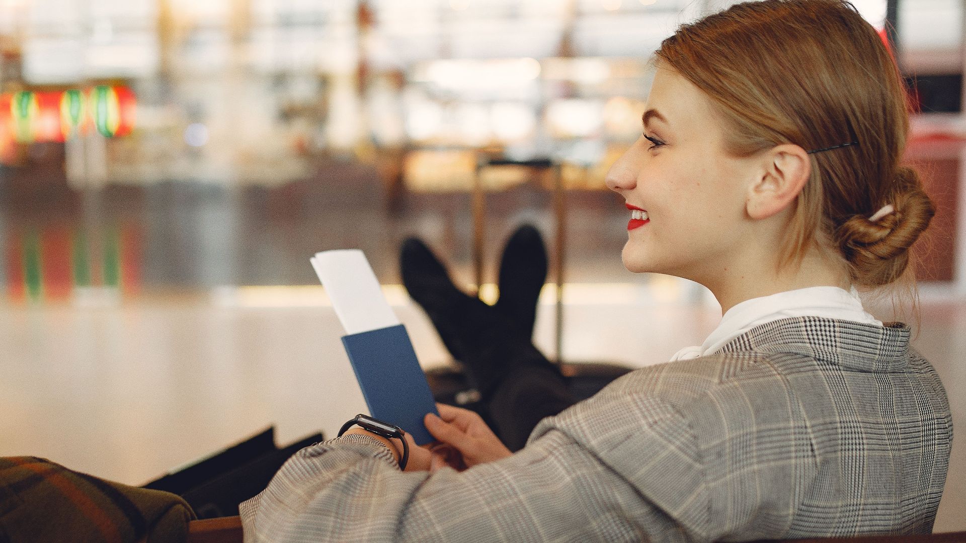 Side view of cheerful female student in checkered jacket smiling away while chilling in hall with outstretched legs and passport in hands