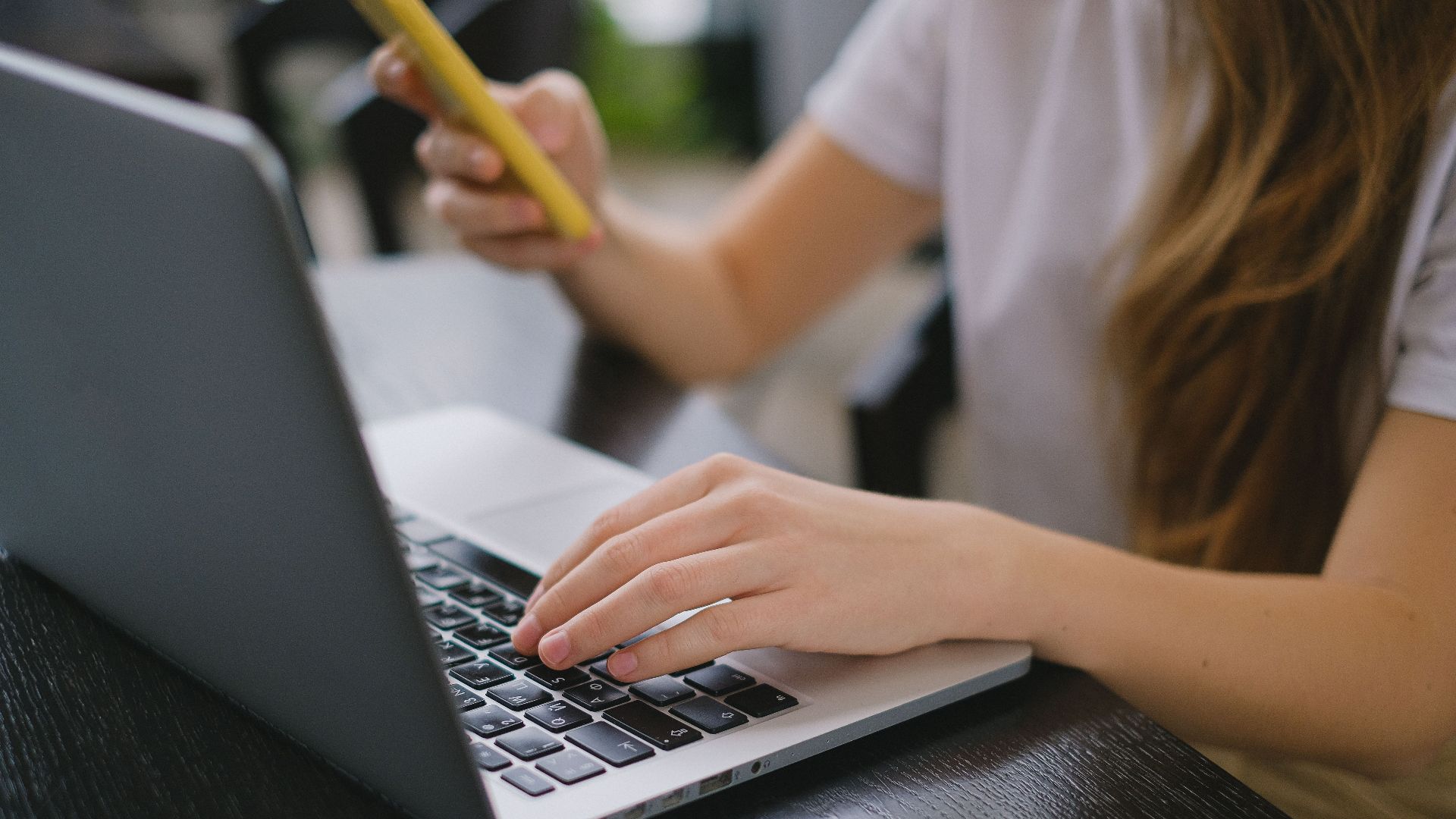 A young woman multitasks by using a laptop and smartphone indoors, focusing on work.