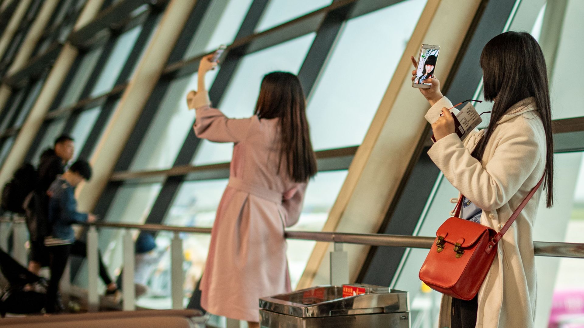 Two young women taking selfies at a modern Shanghai airport terminal with large windows.