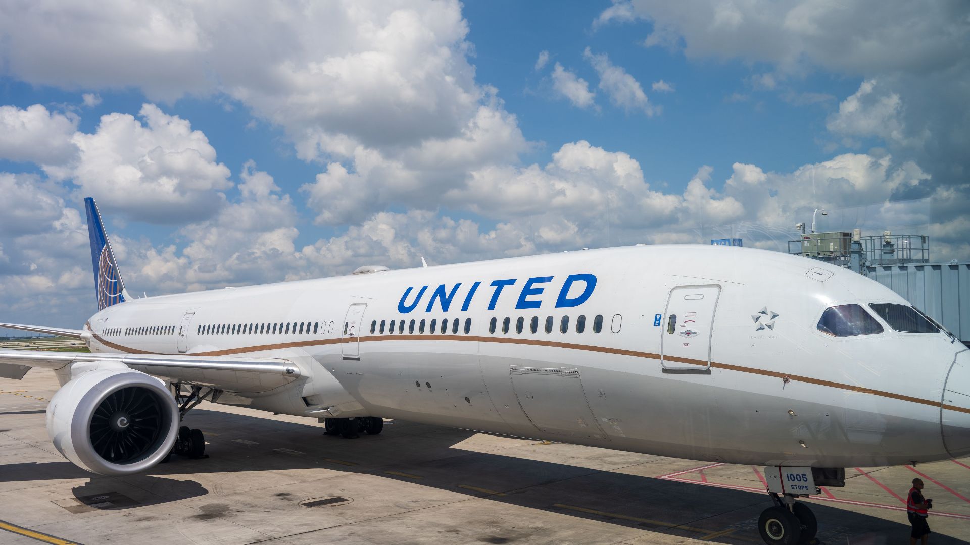 A United Airlines Boeing 787 aircraft parked at the airport terminal under a bright blue sky with clouds.
