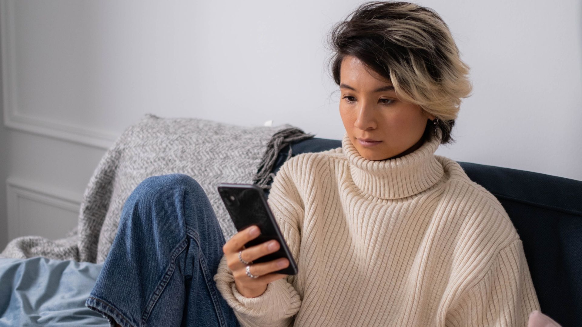Asian woman in cozy sweater using smartphone and laptop on sofa indoors.