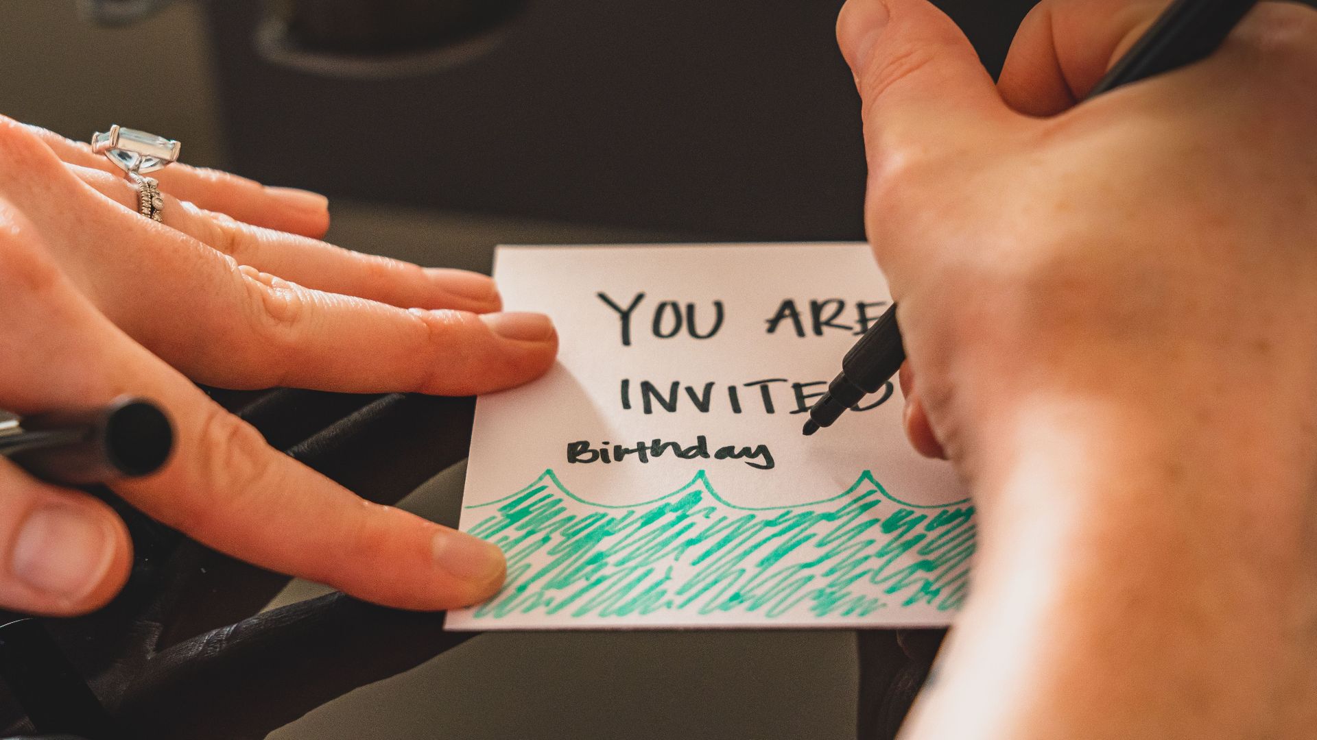 Hands writing a personalized birthday invitation card using a marker on paper.