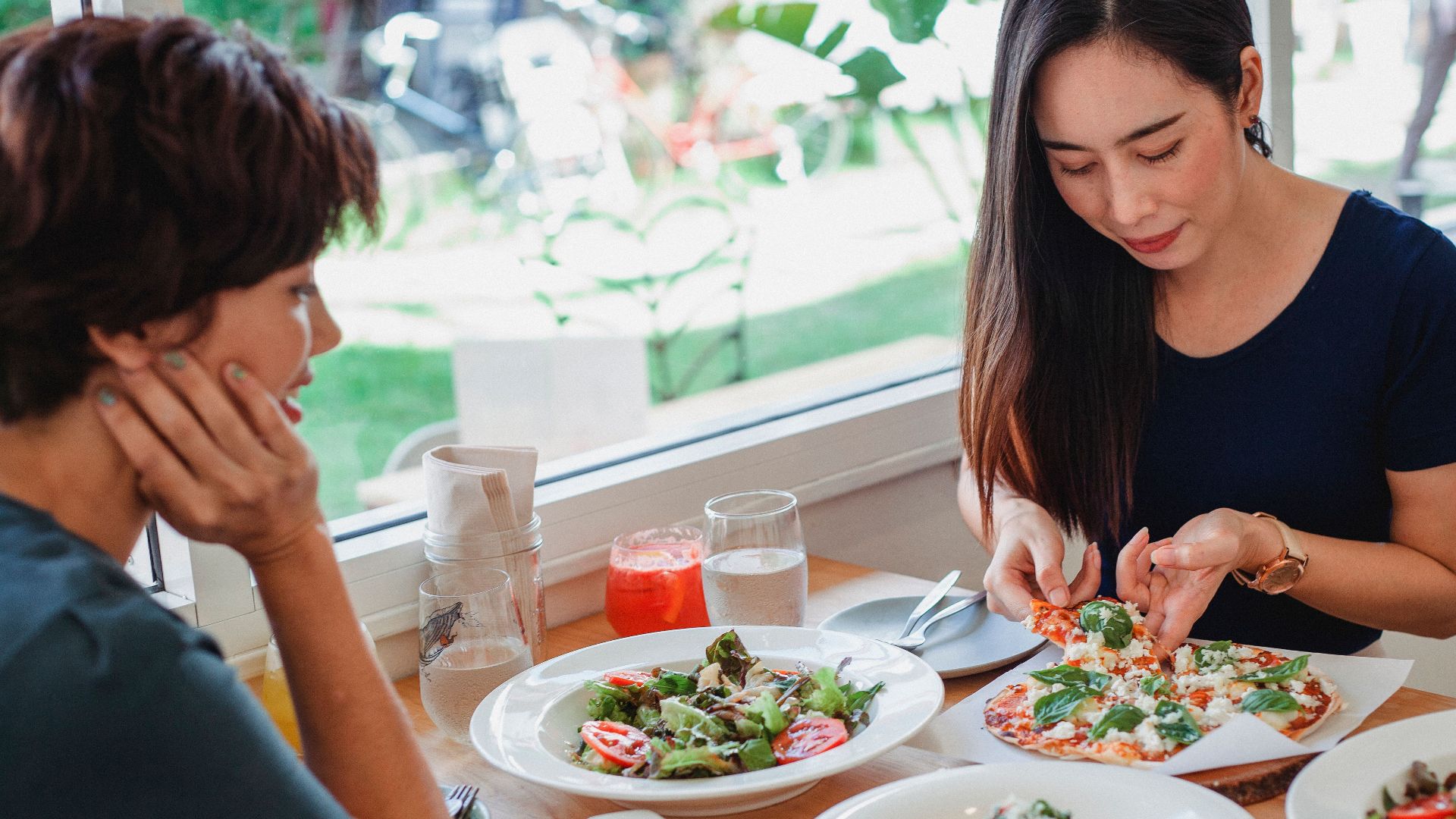 Content Asian woman taking slice of pizza while sitting at table near friend with delicious salad and pasta in restaurant