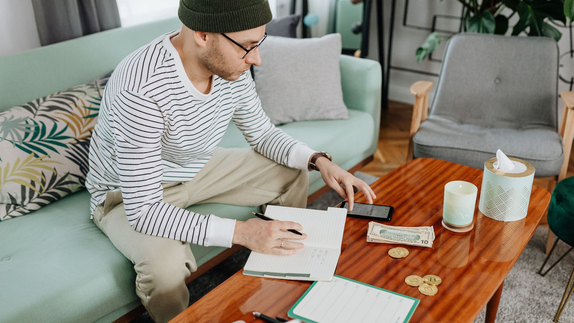 A man in casual attire managing finances with a laptop and notebook on a wooden table indoors.