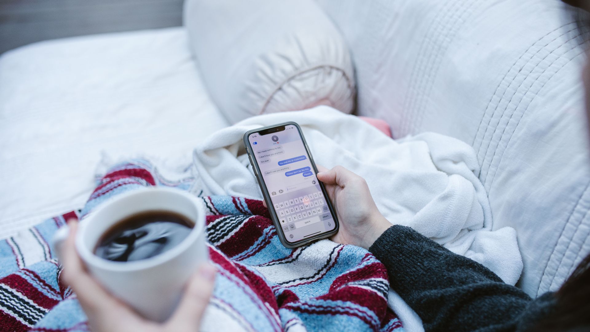 Person relaxing indoors with coffee and smartphone, texting in a cozy setting.