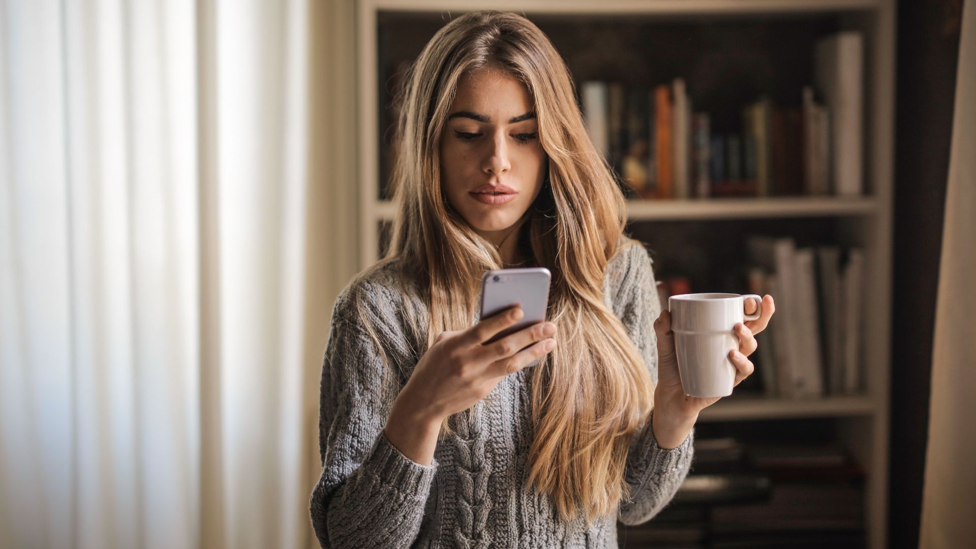 Young woman in a knit sweater texting on a smartphone while sipping coffee at home.