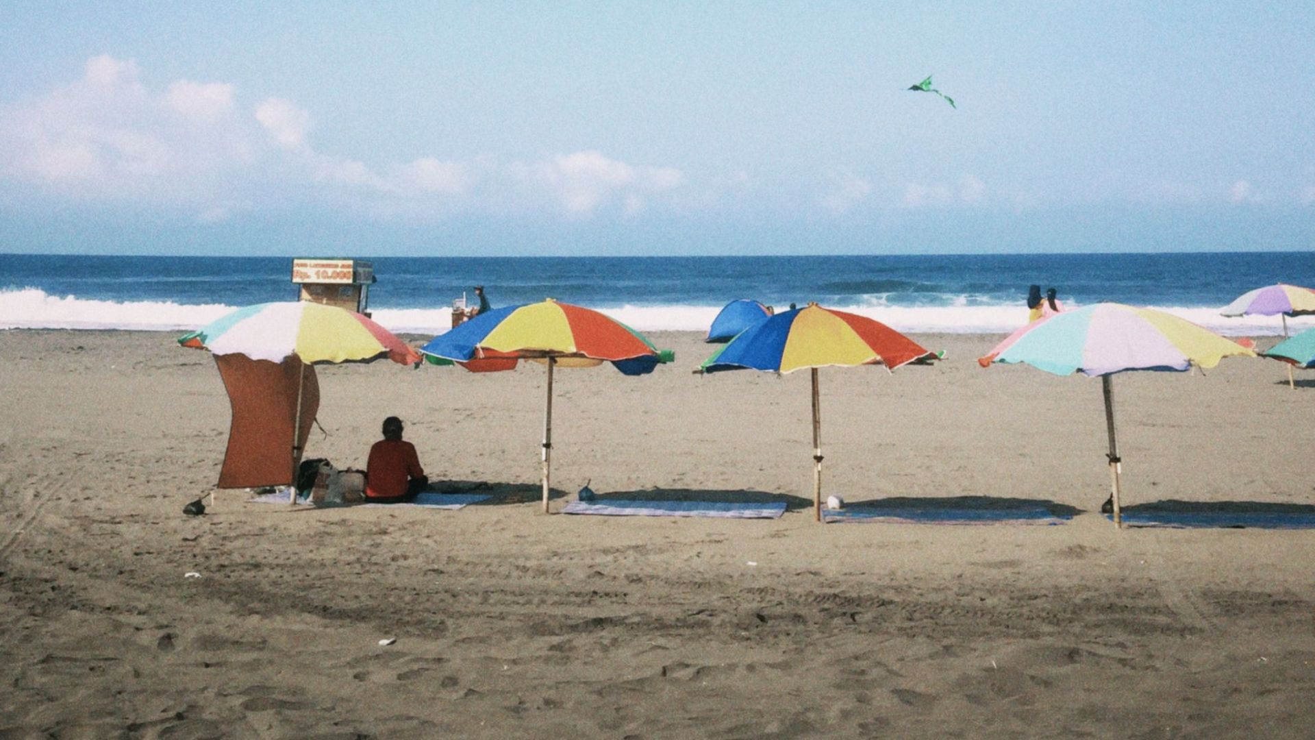 Vibrant beach umbrellas on a sandy shore under a clear blue sky with a distant kite flying.