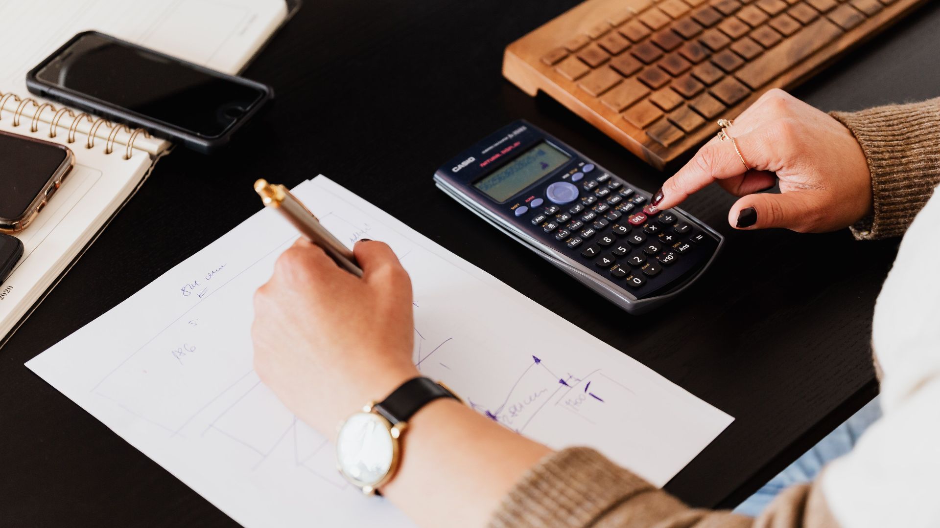 From above crop unrecognizable female accountant wearing stylish watches using calculator and writing down information on paper while working at desk with gadgets