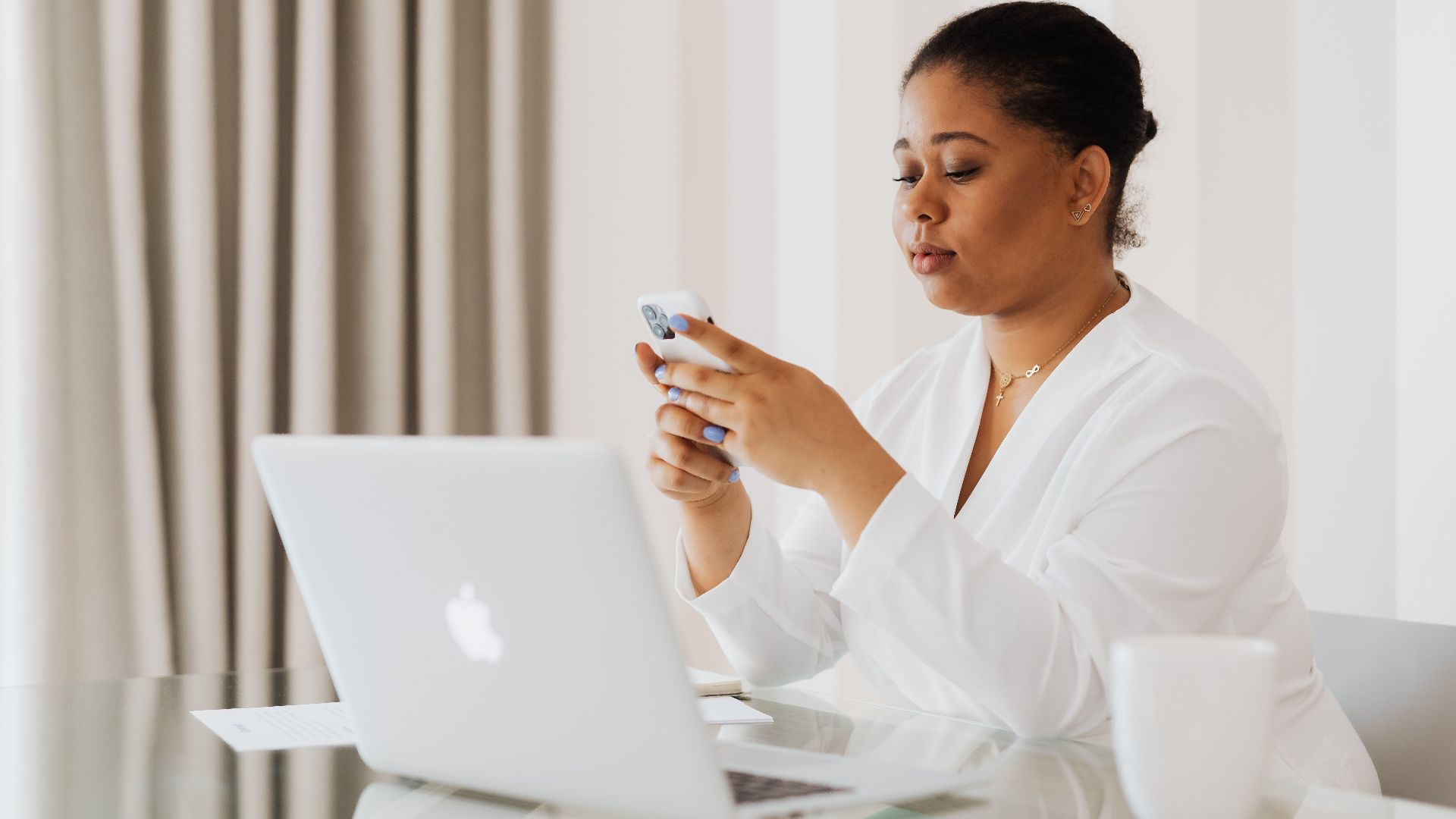 A woman in white attire uses her smartphone while working on a laptop at a desk indoors.