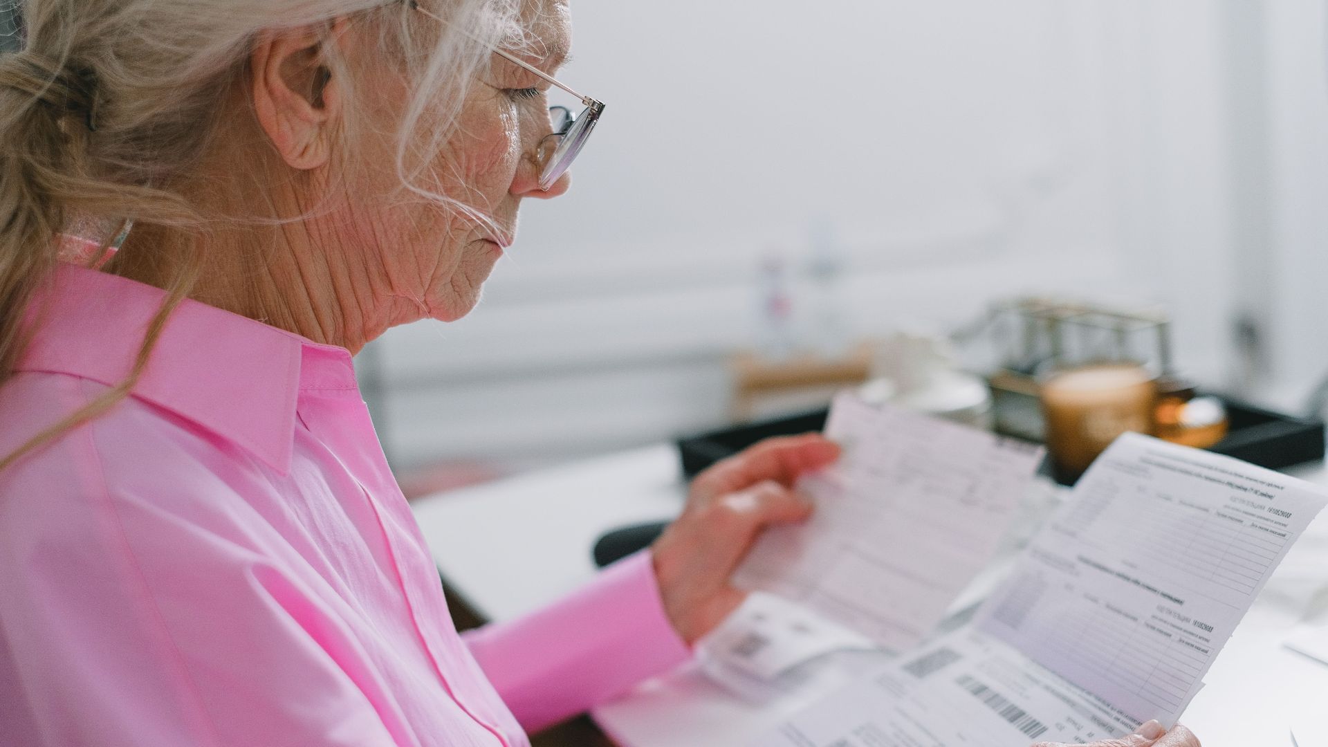 Elderly woman in a pink shirt reviewing paperwork at home.