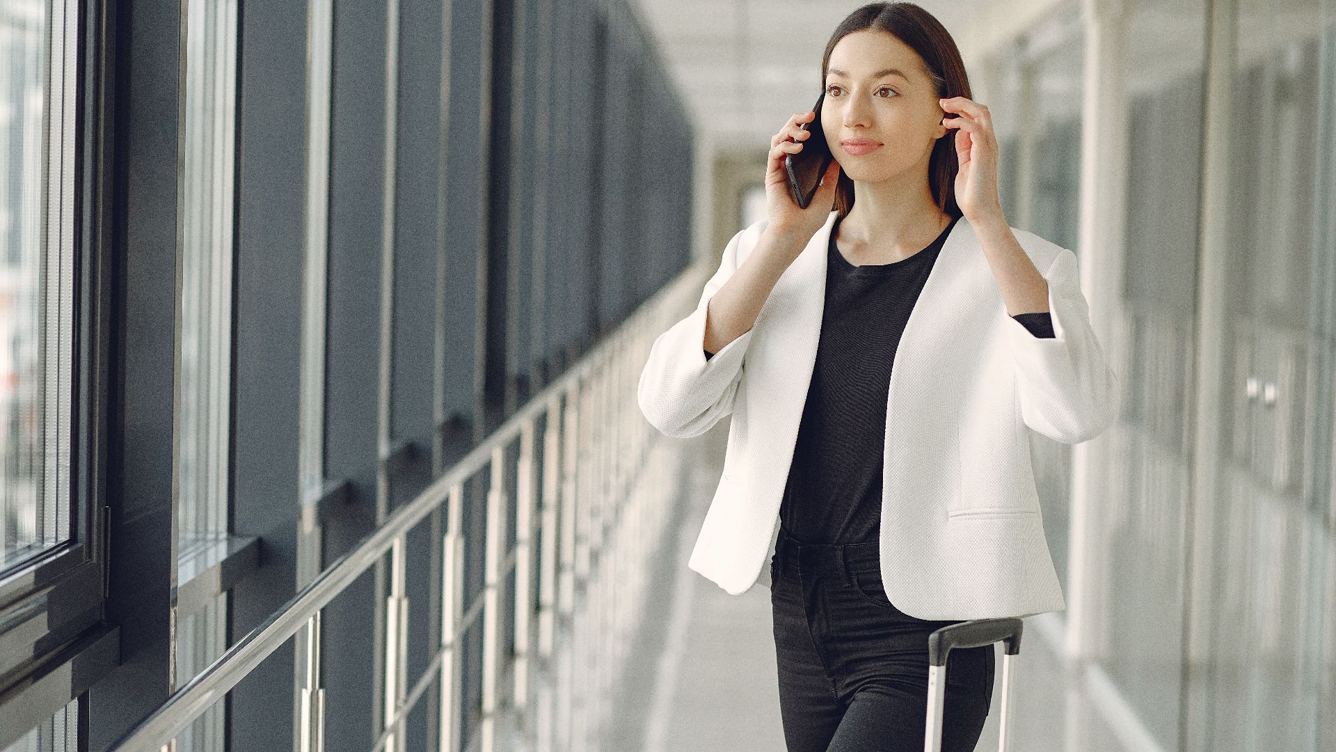 Positive young businesswoman in stylish formal wear with suitcase talking on smartphone and standing in modern airport corridor while waiting for flight