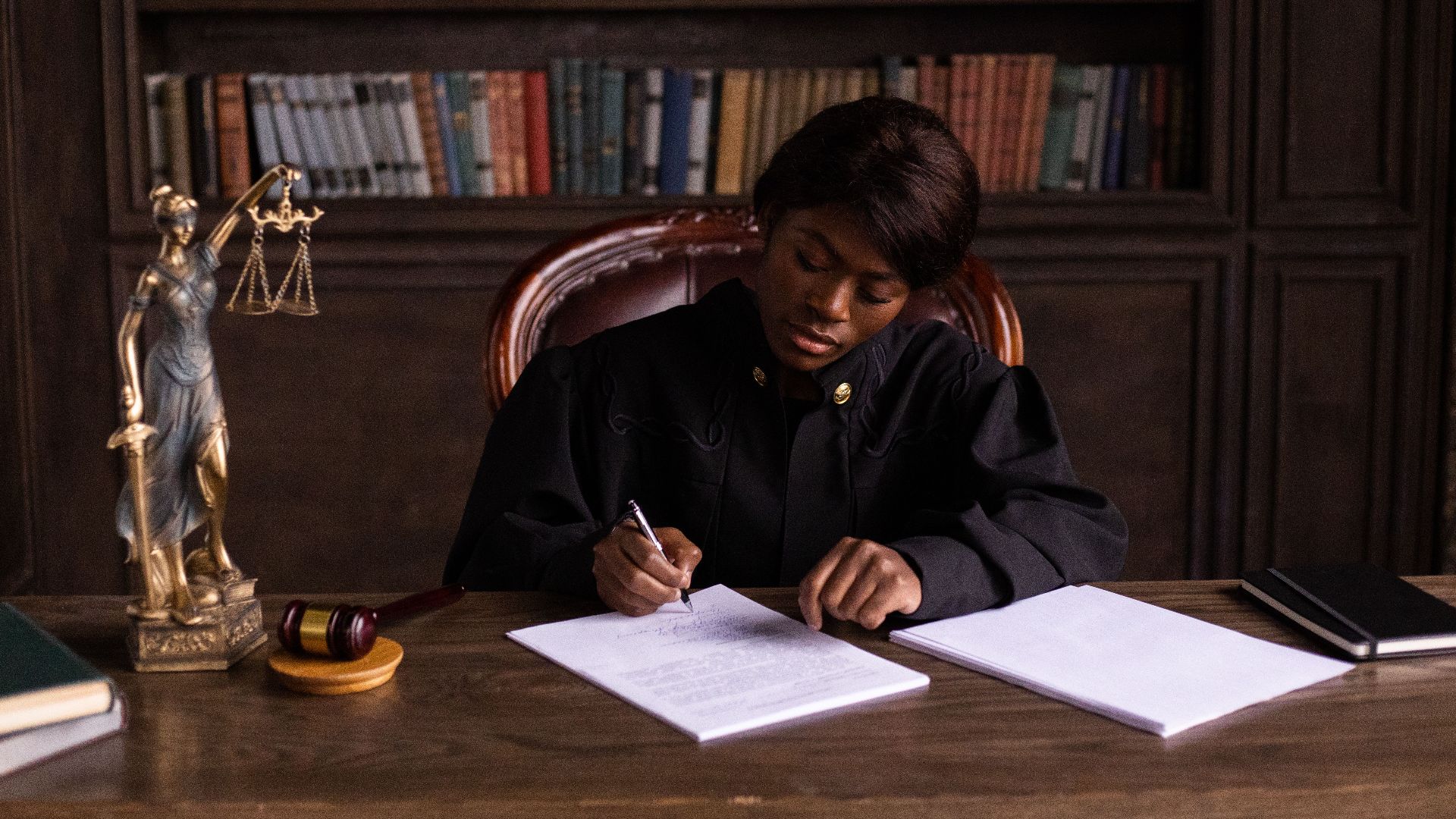 A judge in robes writing on a document at a desk in an office library with law books.