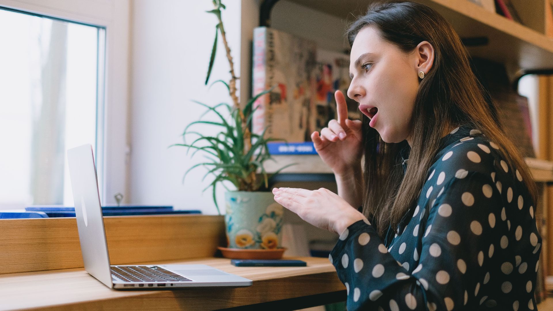 Side view of concentrated young female with long brown hair sitting at table with laptop while practicing sign language during distant lesson at home
