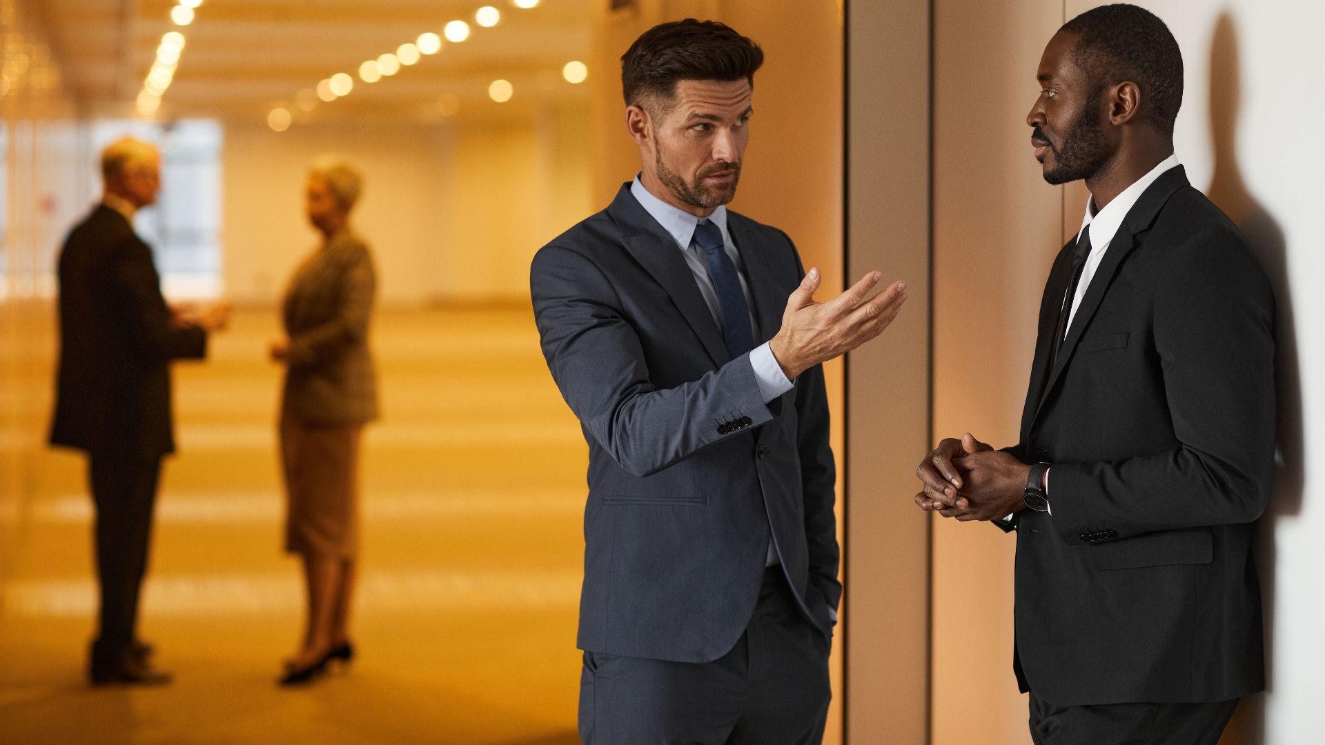 Two businessmen in formal attire engaged in a serious discussion in a modern office hallway.