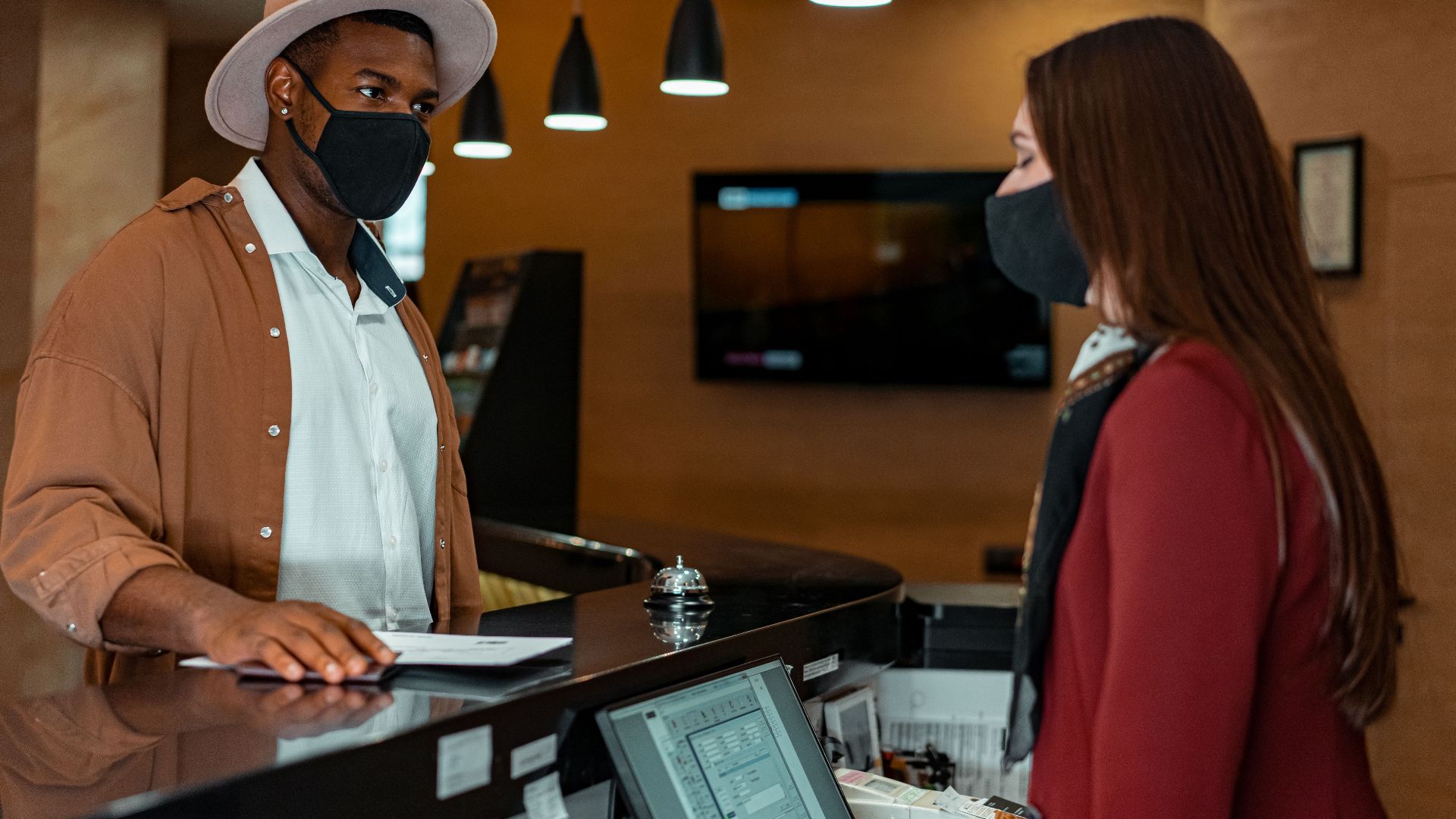A man and woman in face masks checking in at a hotel reception, emphasizing safety protocols.