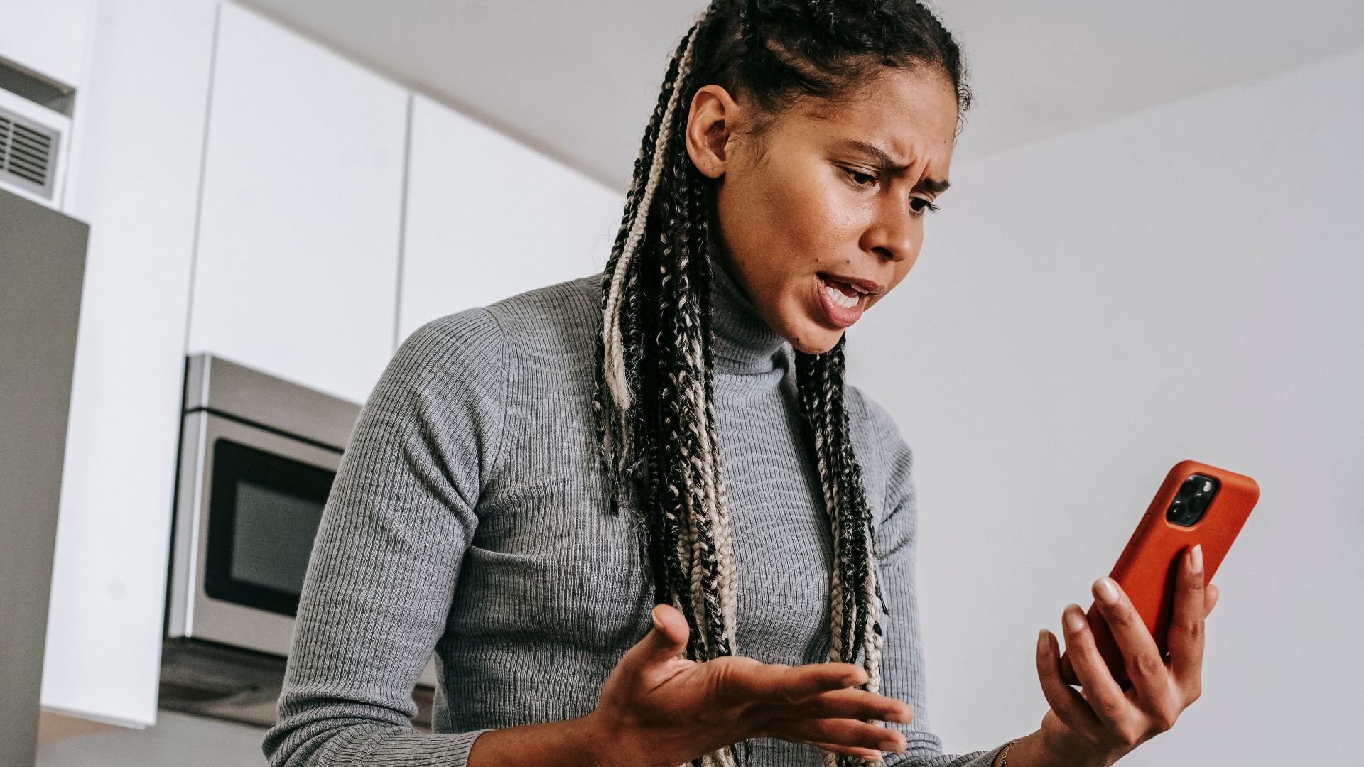 From below of angered black female with Afro braids in casual outfit gesticulating and resenting while having video call on smartphone in modern light kitchen