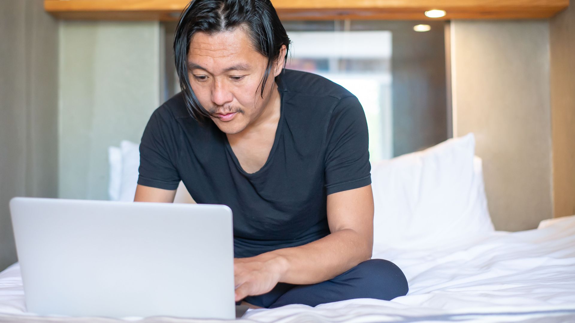 Asian man working remotely on a laptop in a cozy bedroom setting, exemplifying modern work-from-home lifestyle.