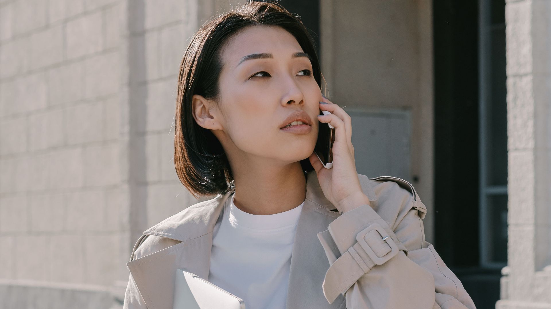 Confident Asian businesswoman in trench coat making a phone call while walking outside an urban building.