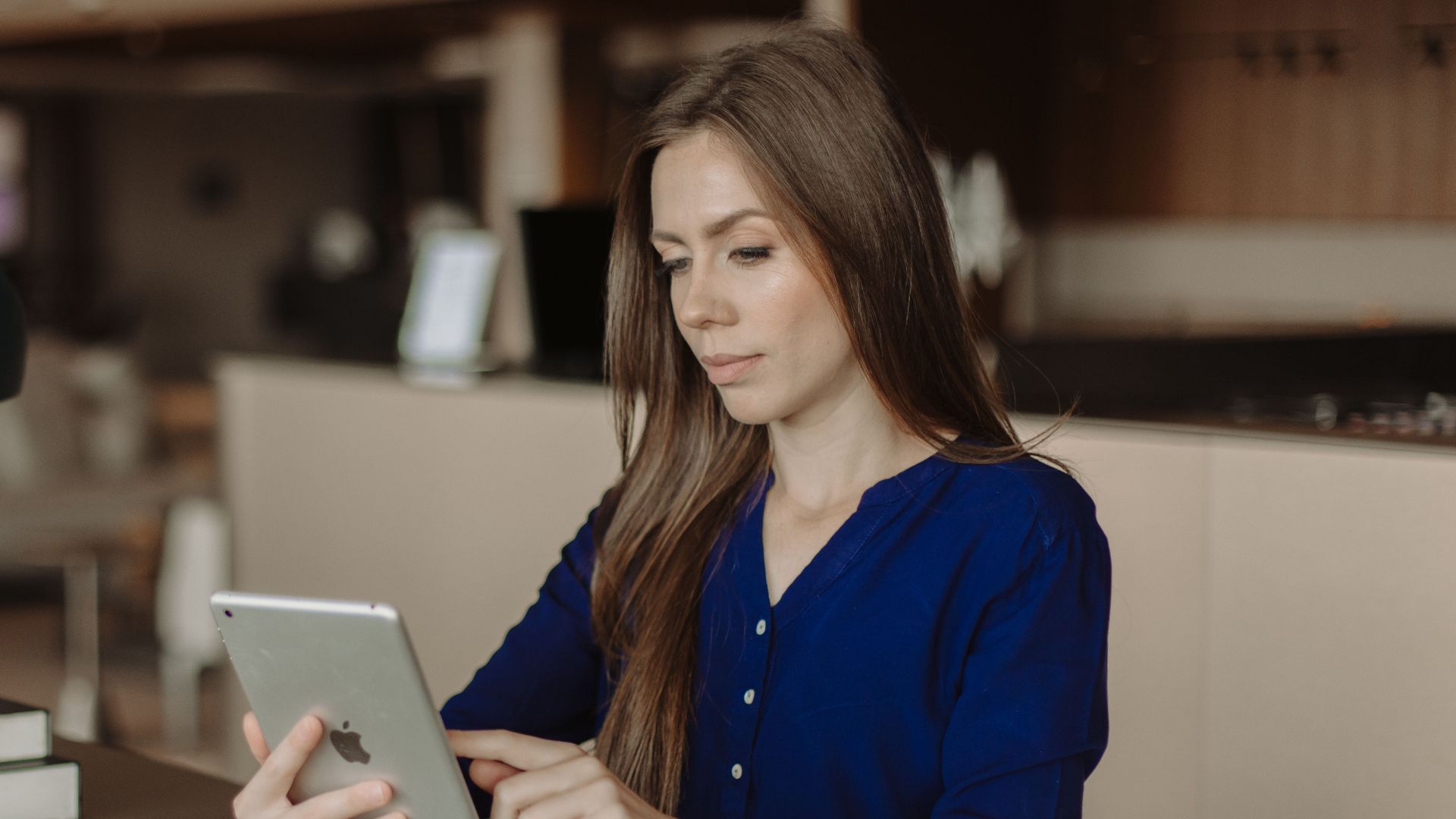 Woman reading on a tablet at a desk with books in a modern office setting.
