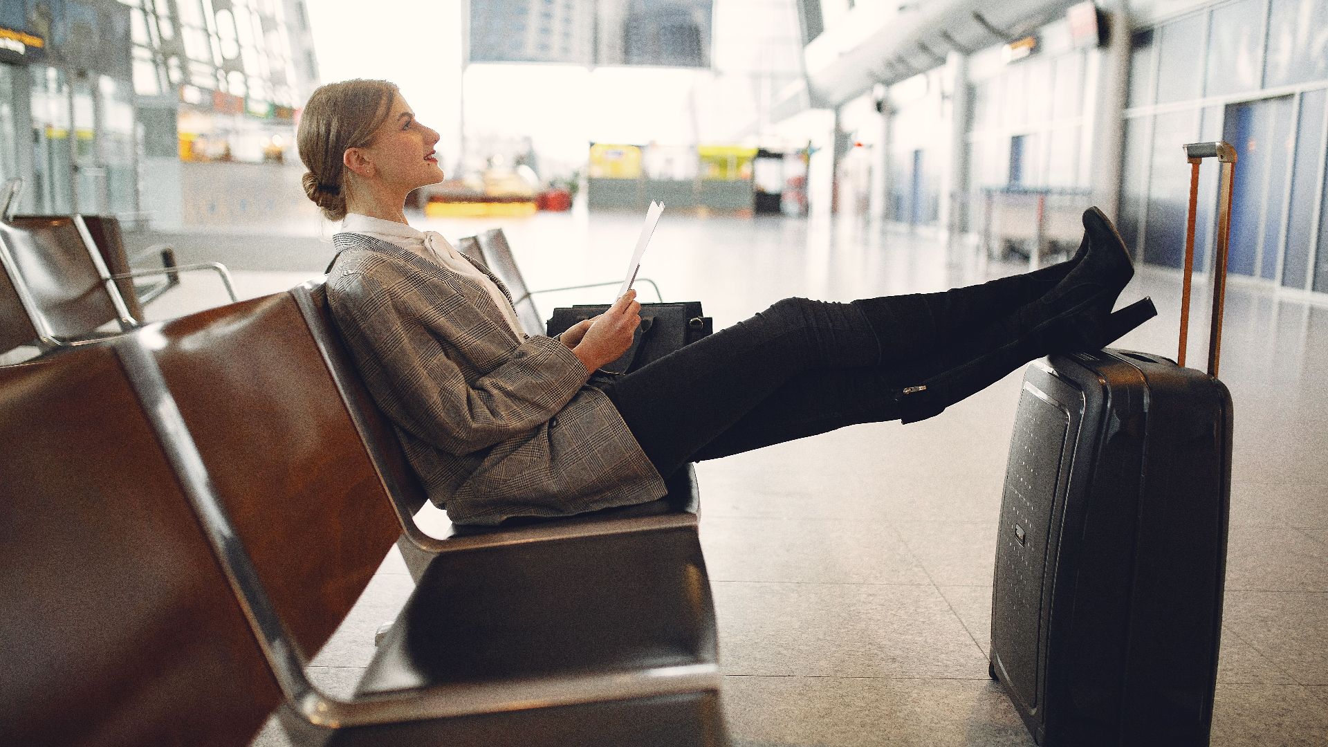 A woman relaxes in an airport terminal waiting area, reading a tablet with luggage nearby.