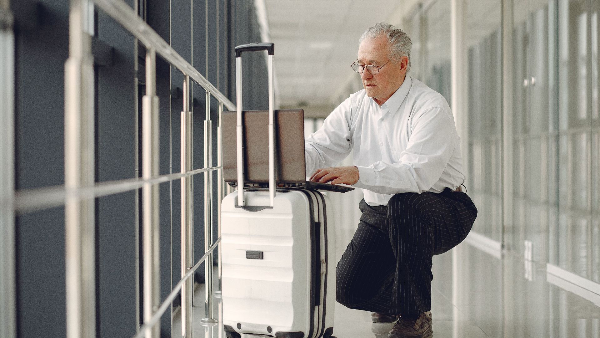 Full length of focused mature male in formal wear using laptop on baggage while having semisitting position in airport corridor