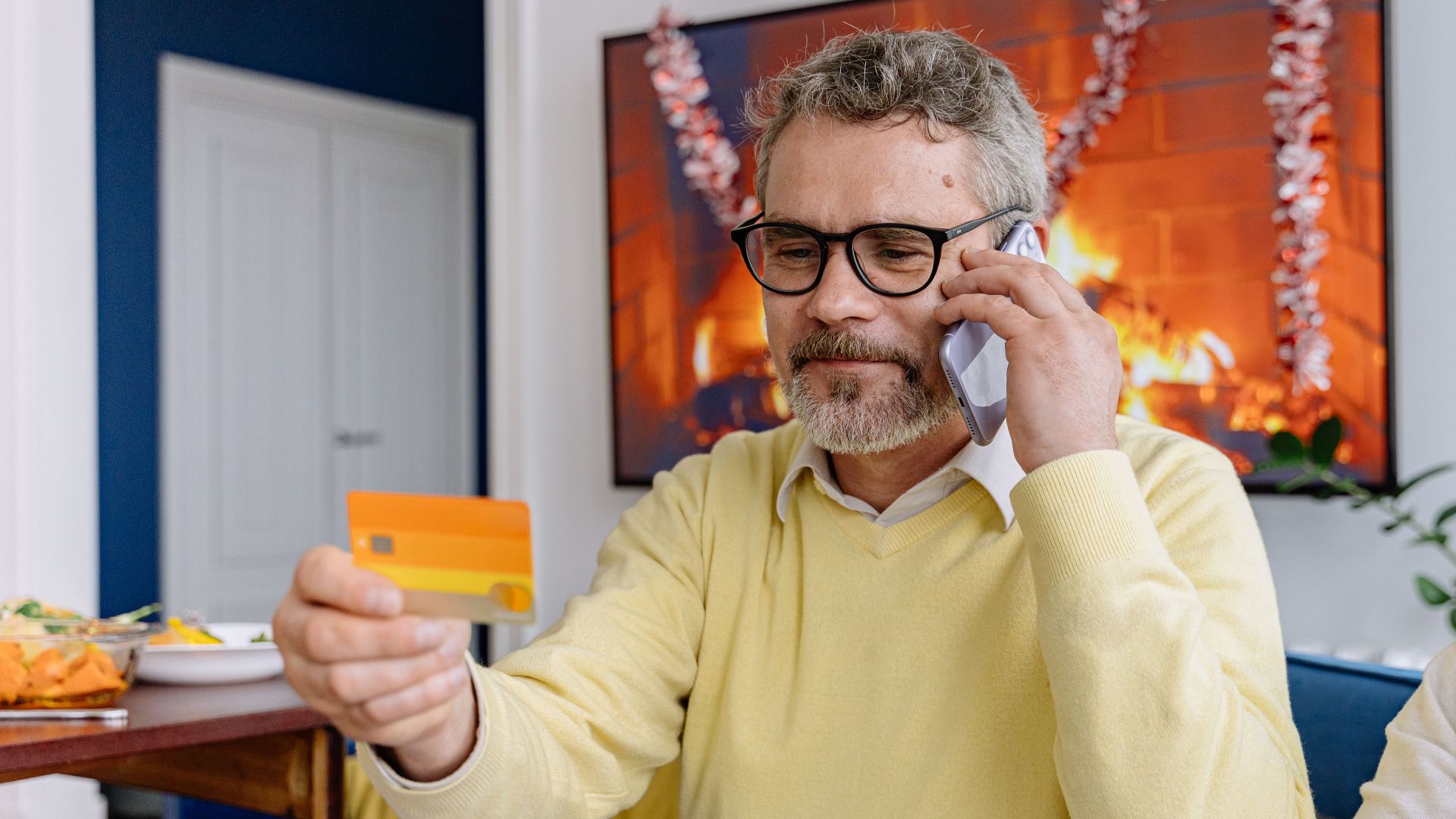 Elderly man using phone while holding a credit card indoors near a cozy fireplace.