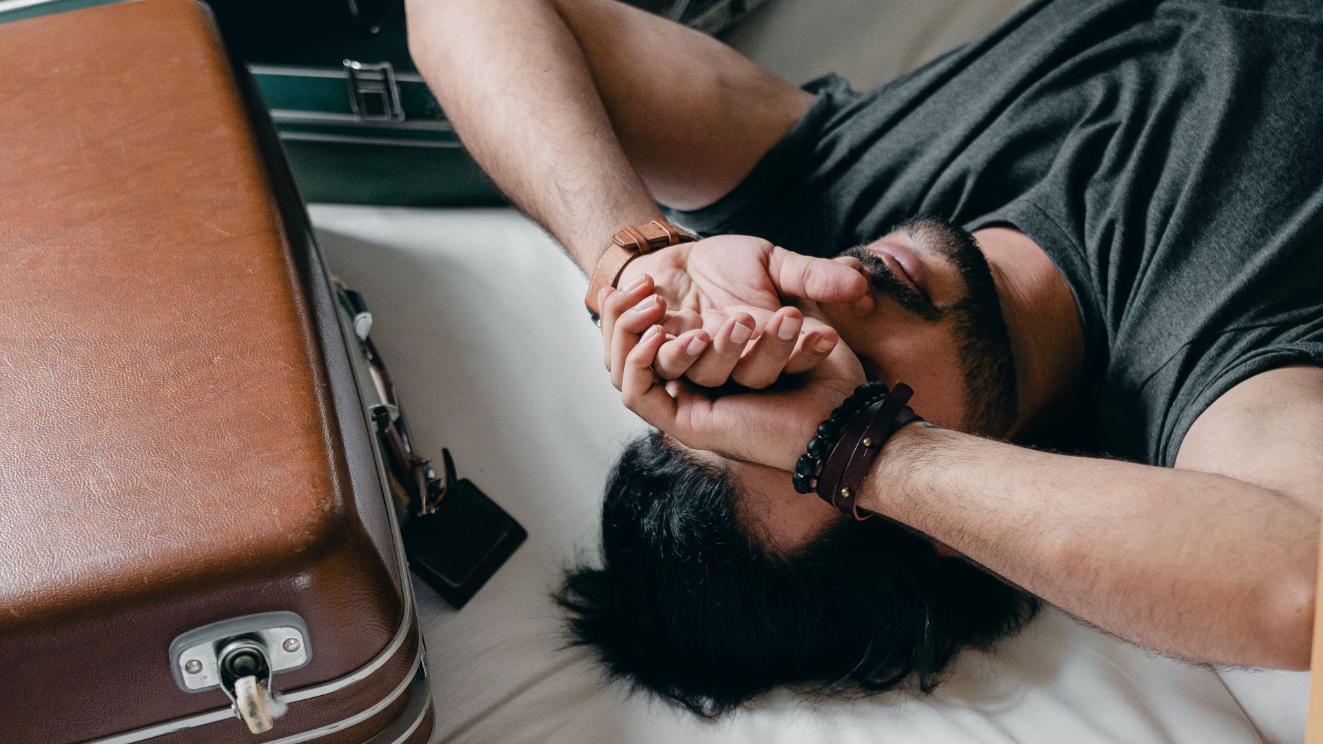 From above young bearded male wearing gray t shirt resting on bed near suitcases and covering eyes with arms while moving into new apartment
