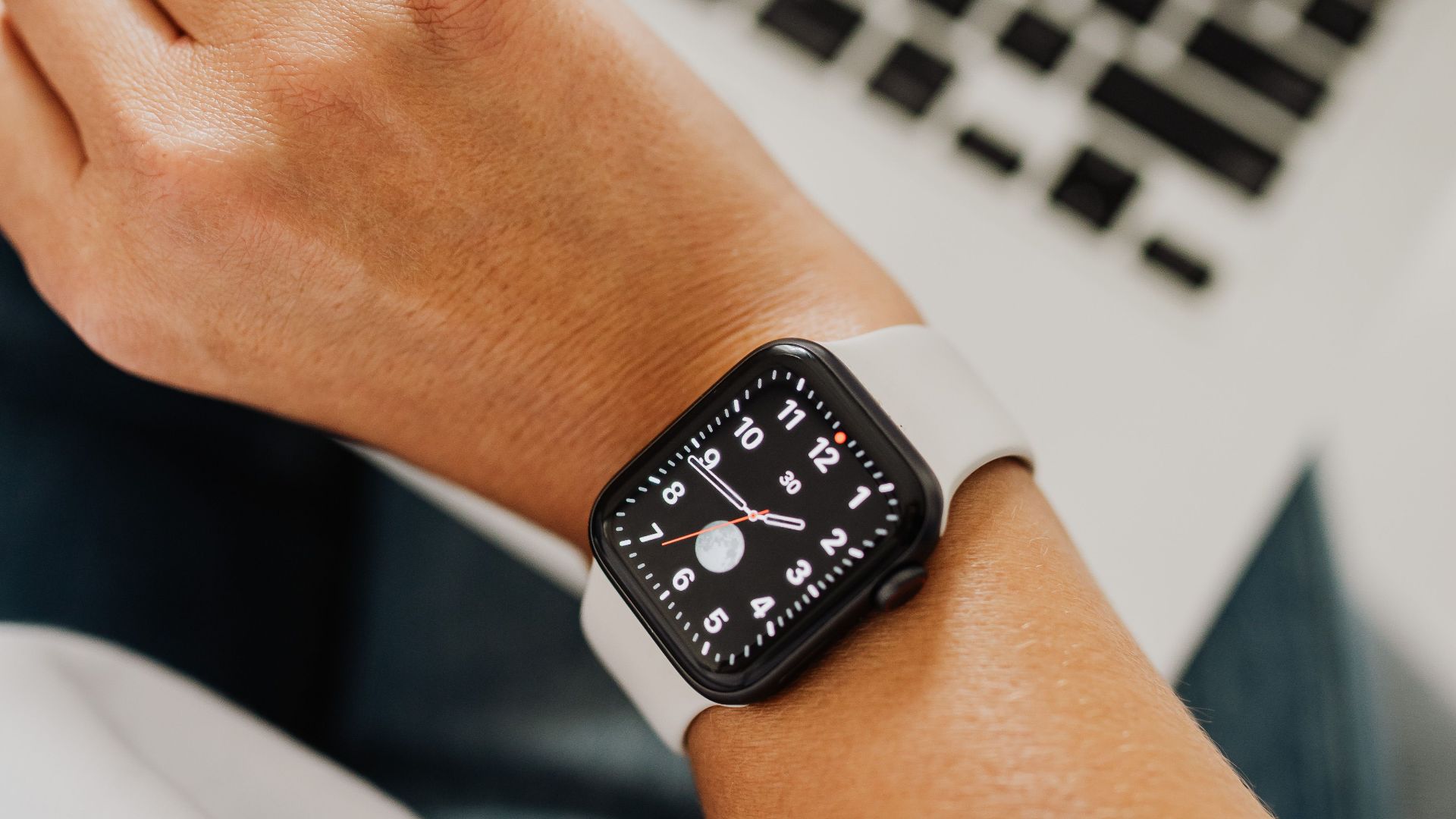 Close-up of a hand wearing a smartwatch positioned next to a laptop in a workspace setting.