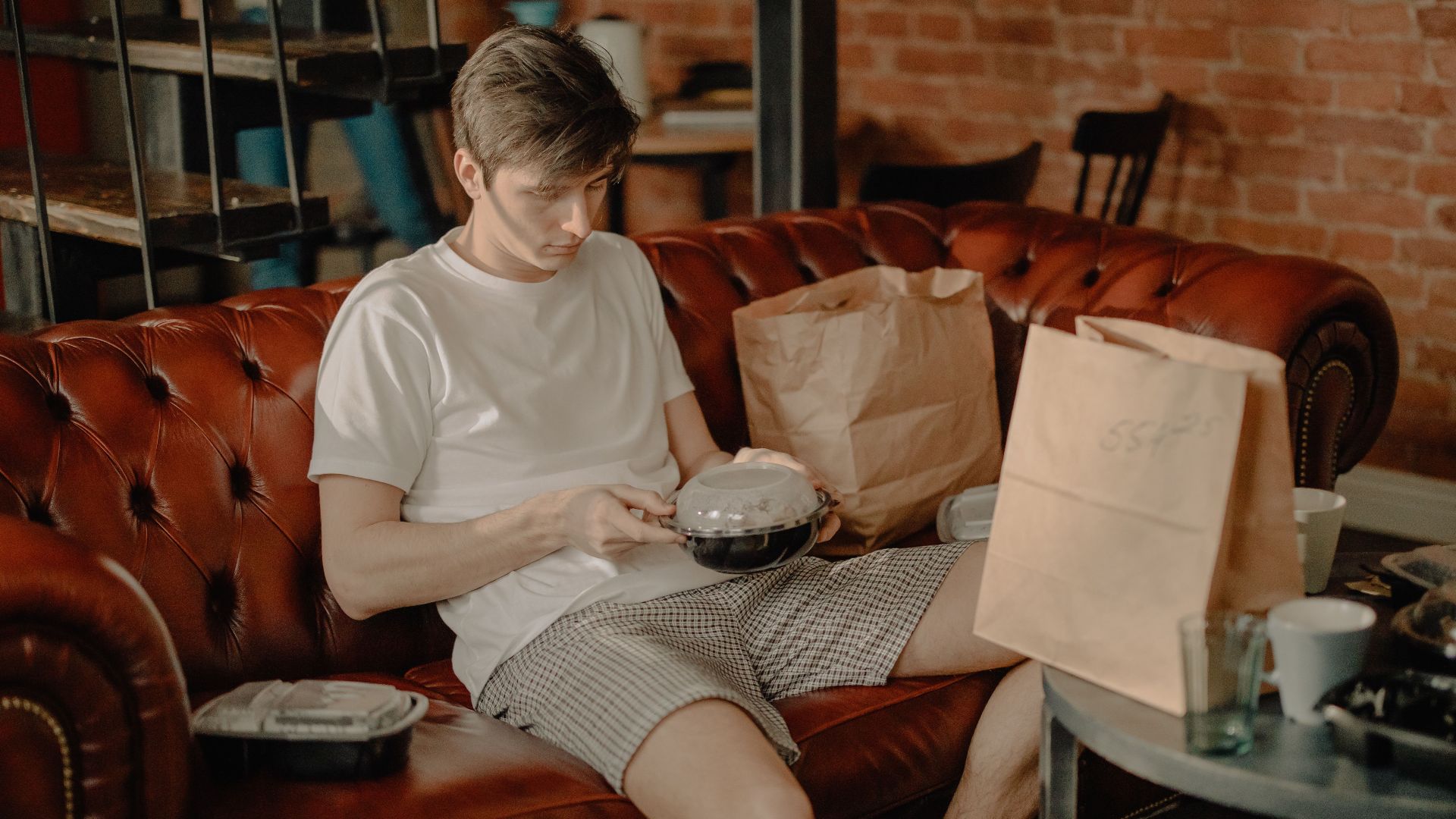 A young man sits on a leather couch enjoying a takeout meal in a cozy home interior.