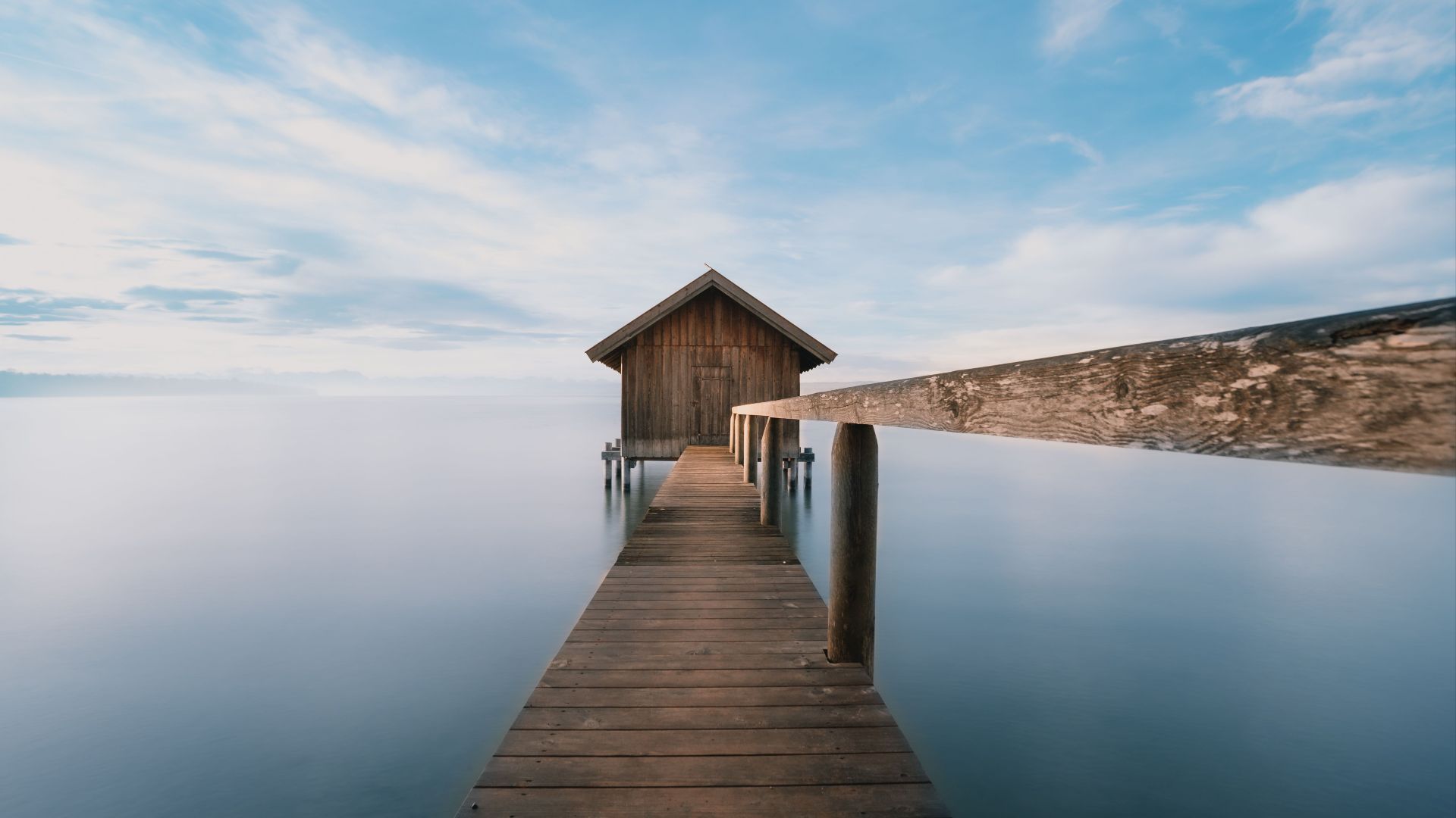 A serene lakeside cabin with a wooden pier extending over calm waters under a soft sky.