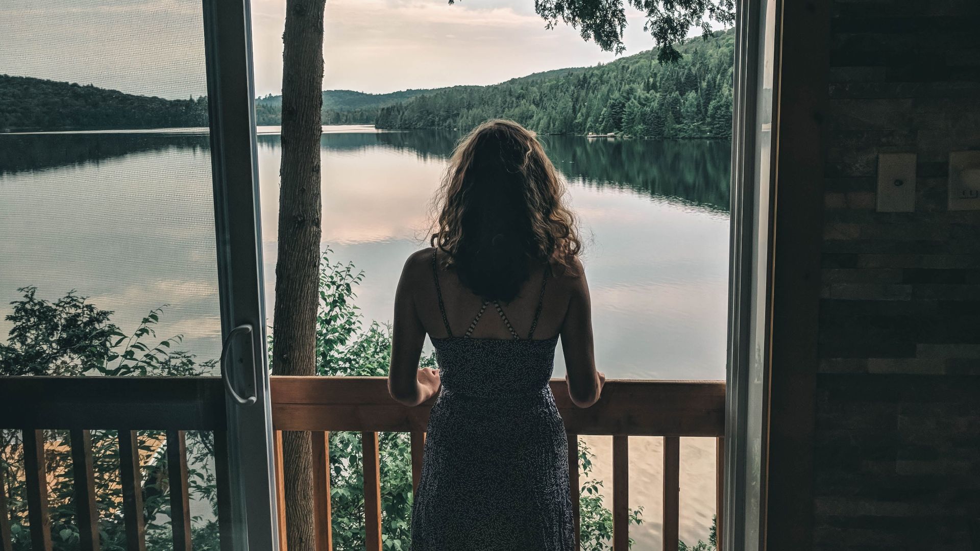 A woman stands on a balcony overlooking a peaceful Canadian lake surrounded by lush greenery.